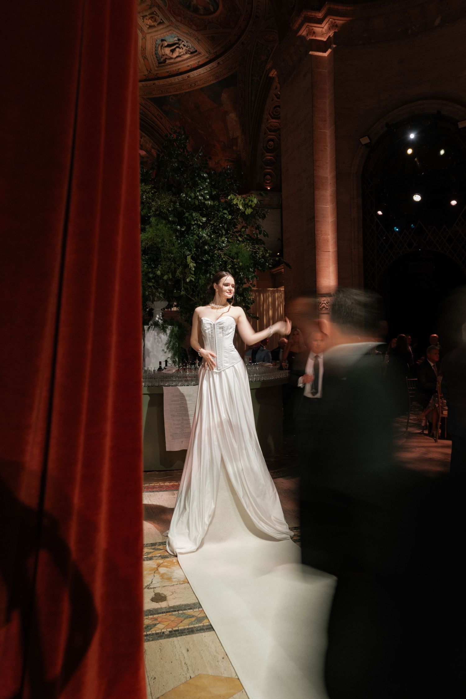 A woman in a white wedding gown standing on a stage with red curtains in the background.