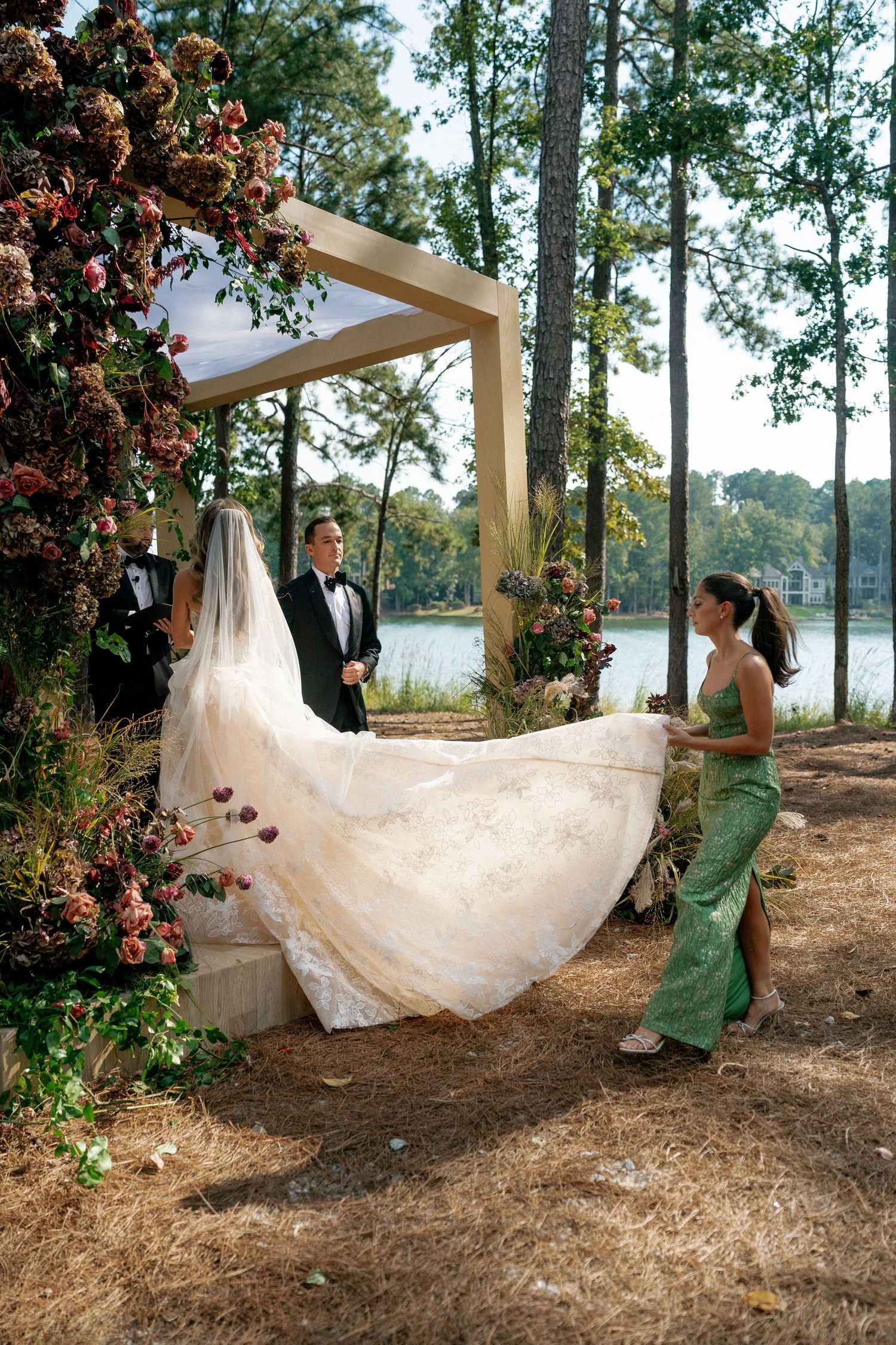 Wedding dress being arranged by assistants under wooden arch with floral decorations.