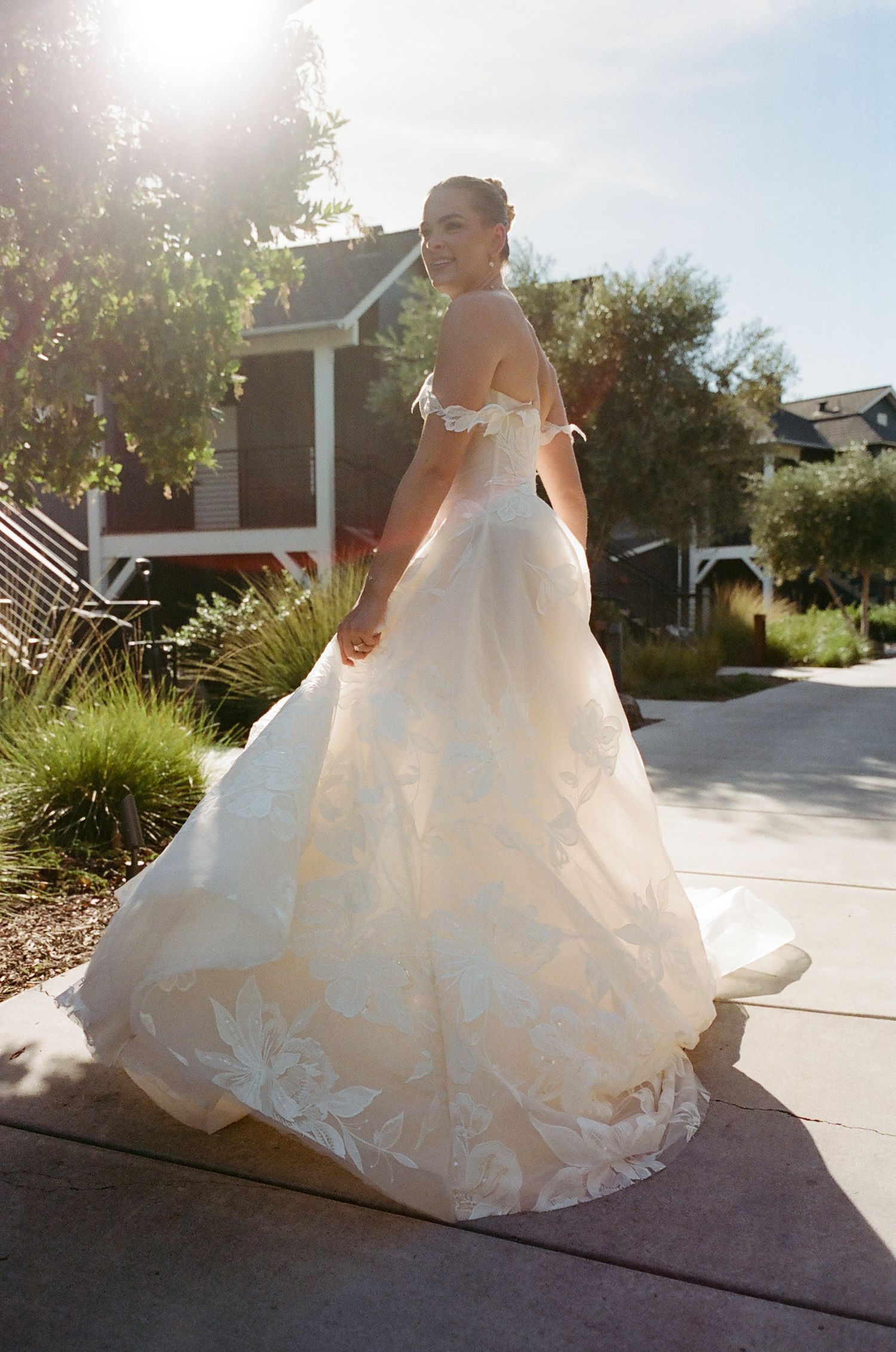 Flowing white wedding gown with intricate lace details trailing on a sunlit pathway.