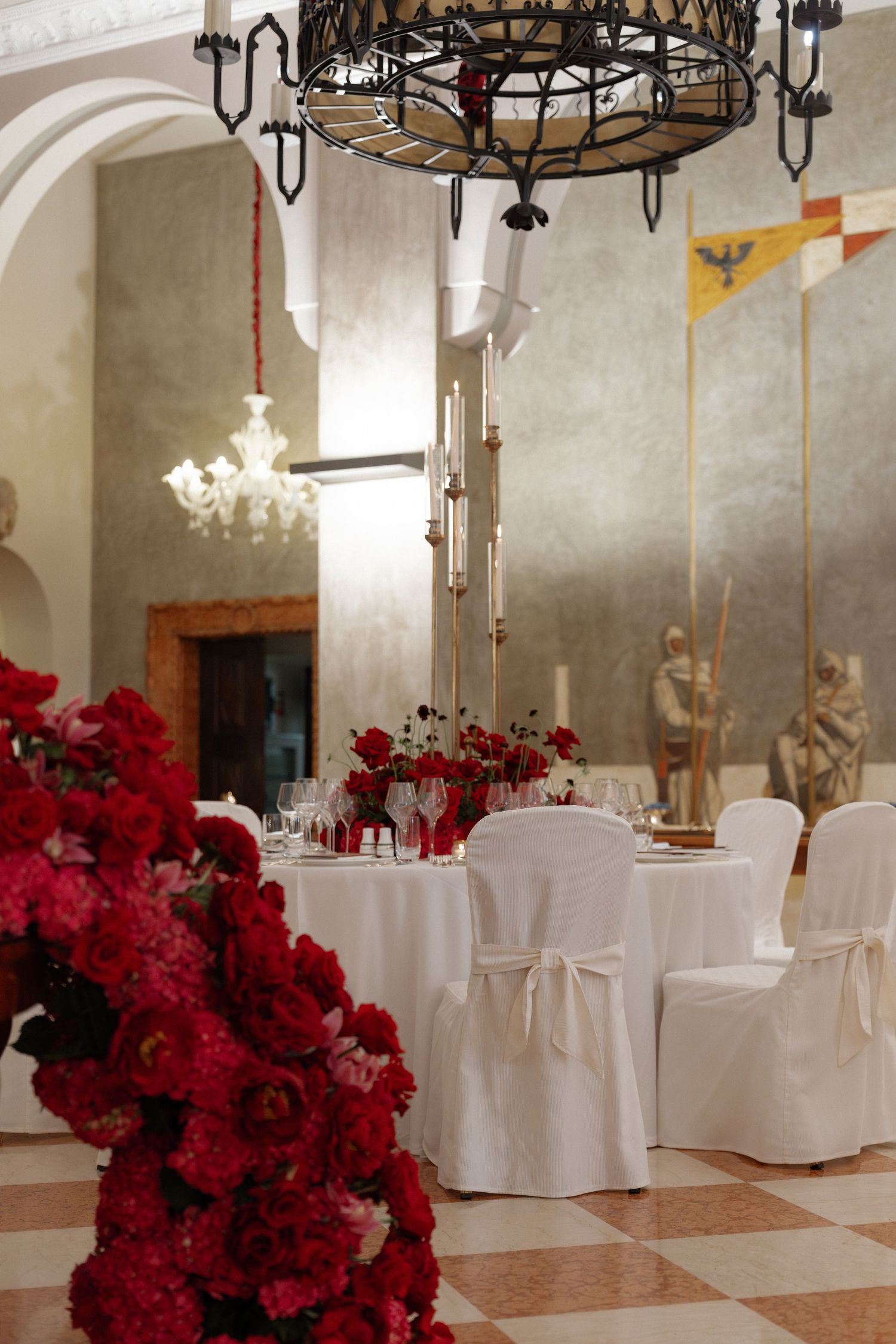 Elegant wedding reception hall with white chair covers, red floral centerpieces, and ornate iron chandelier.