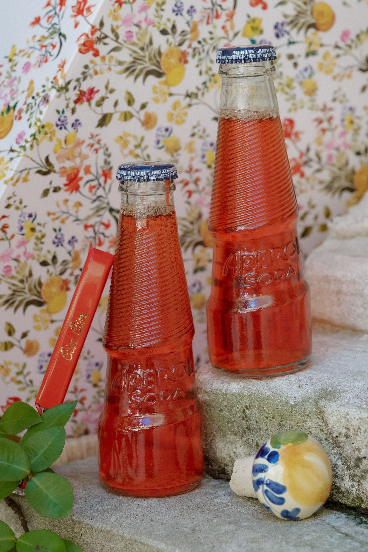 Two vintage glass bottles filled with red liquid on stone steps, with a floral wallpaper background.