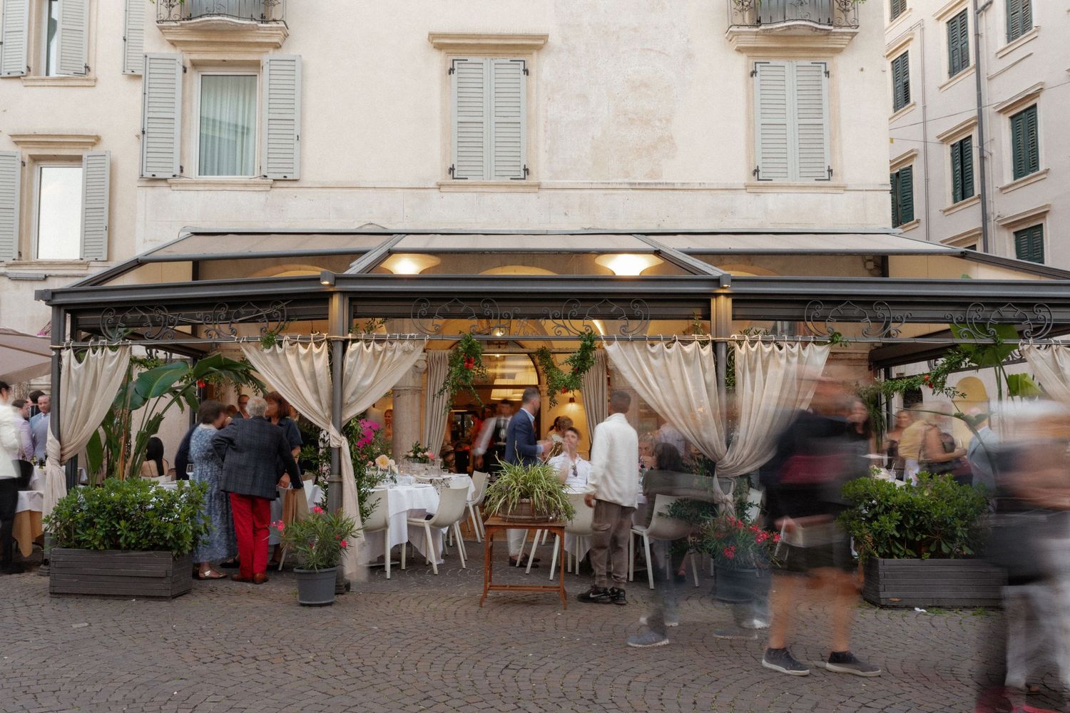 Outdoor restaurant terrace with elegant curtains and lush greenery on a cobblestone piazza with guests dining.