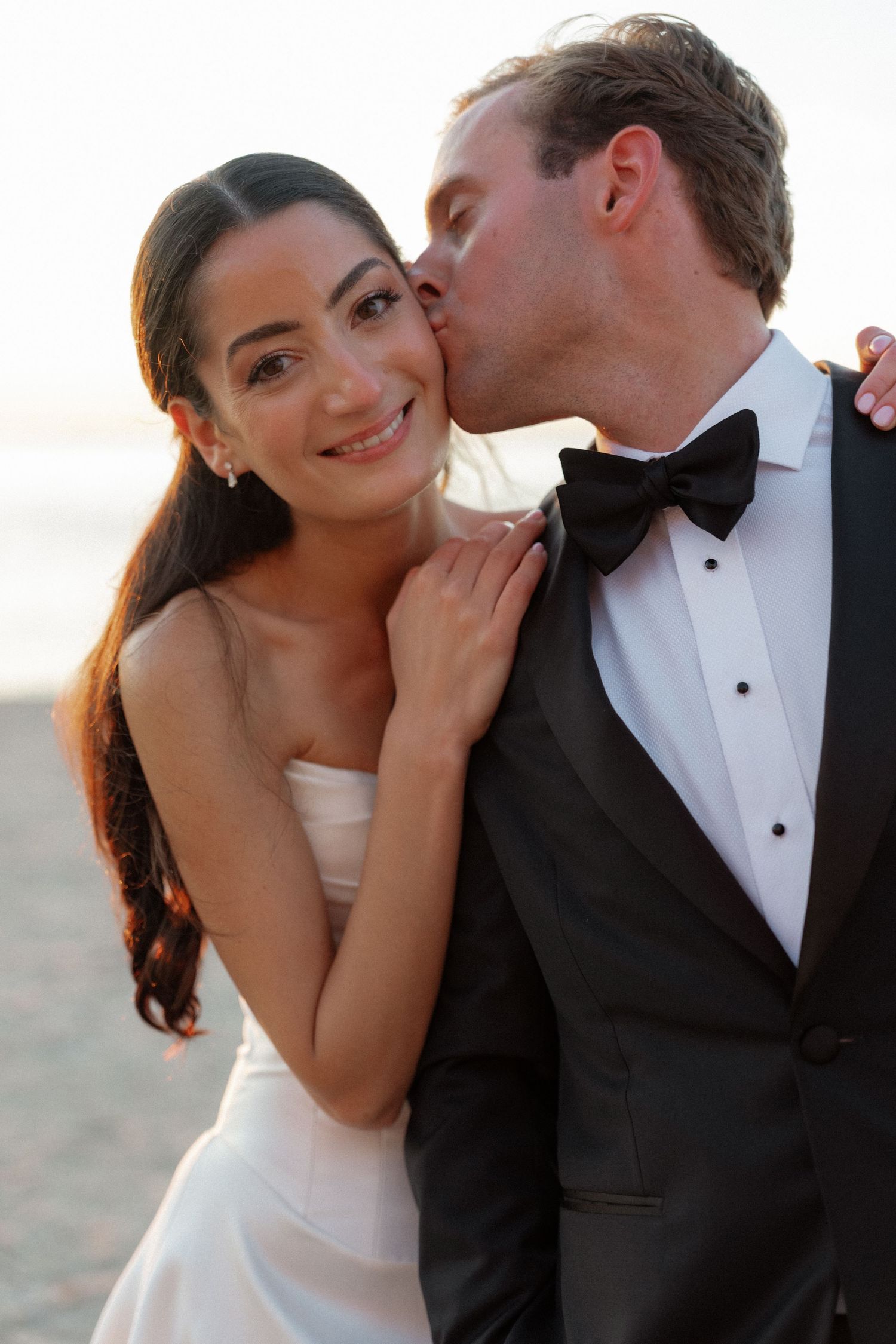 Groom in black tuxedo kissing smiling bride on cheek at sunset.