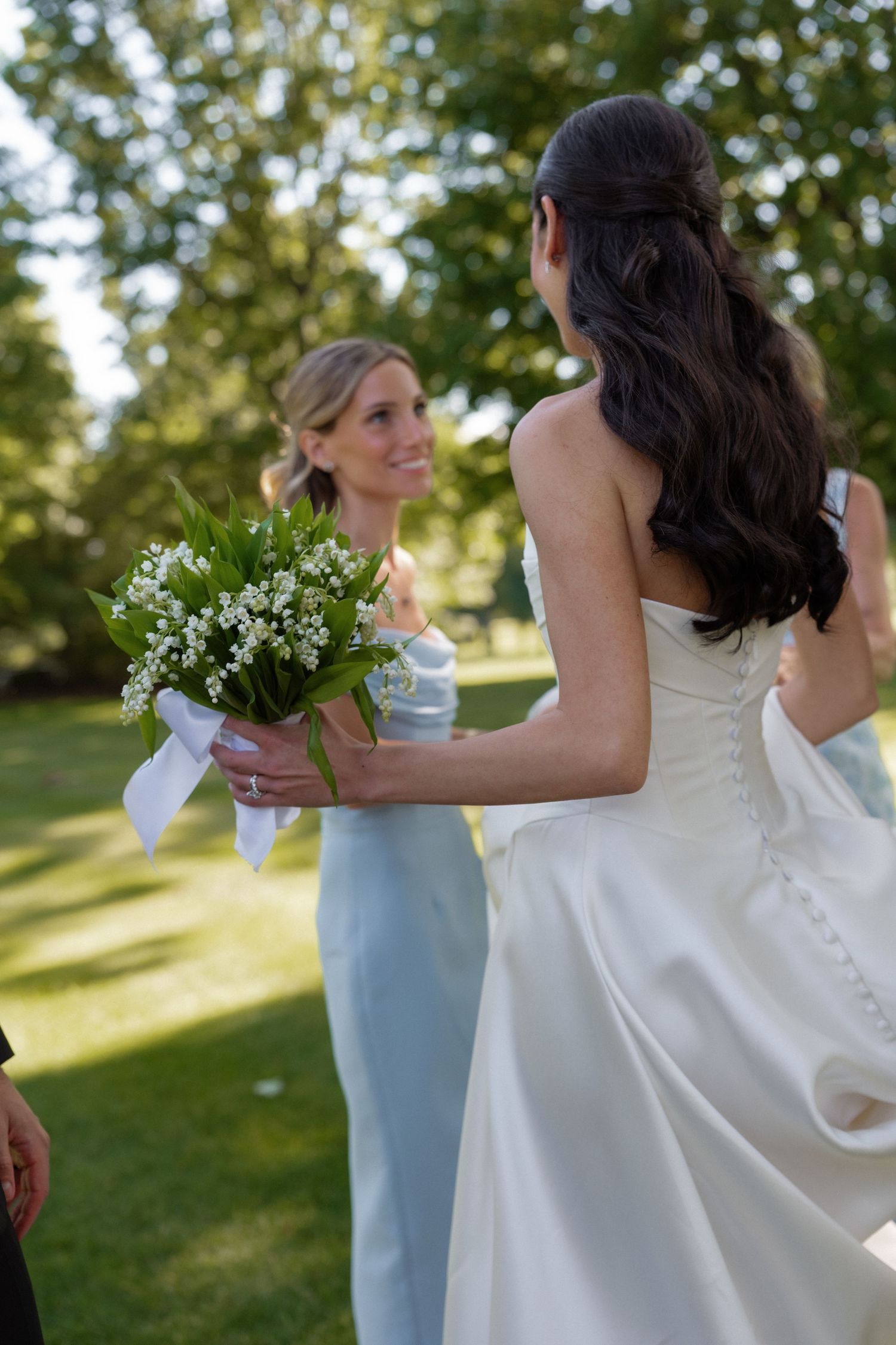 Bride holding lily of the valley bouquet turns to smile at bridesmaid in a sunlit garden.