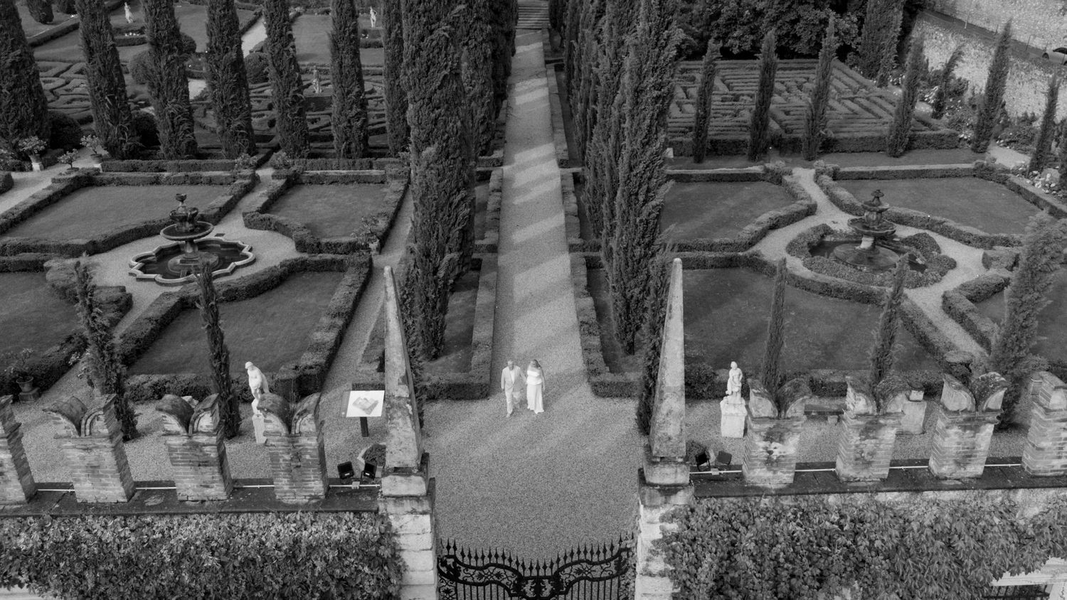 Aerial black and white photo of a formal garden with symmetrical hedges, fountains, statues, and two figures on a central path.