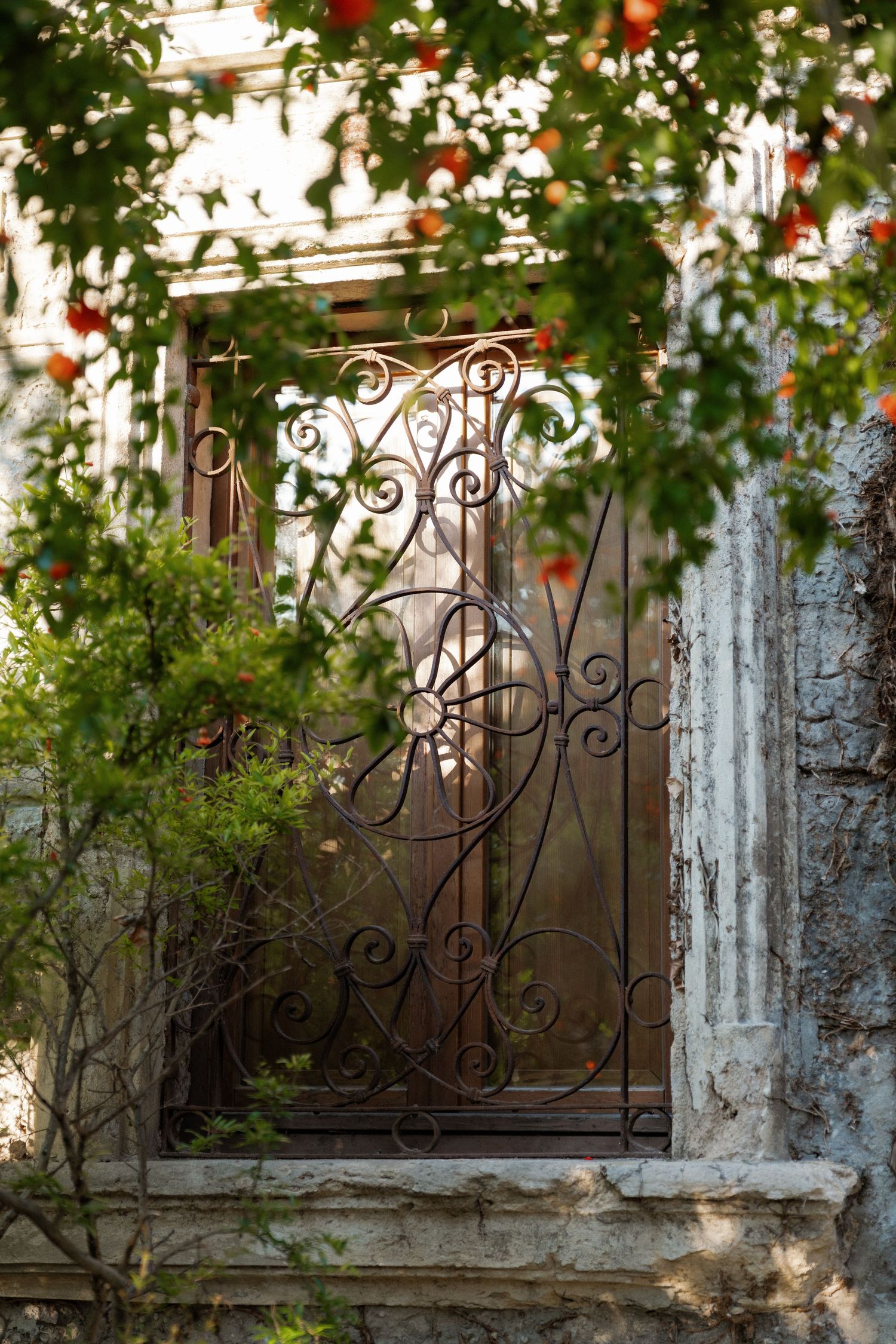 Ornate rusty wrought iron gate surrounded by lush green foliage and red flowers at an old stone building.