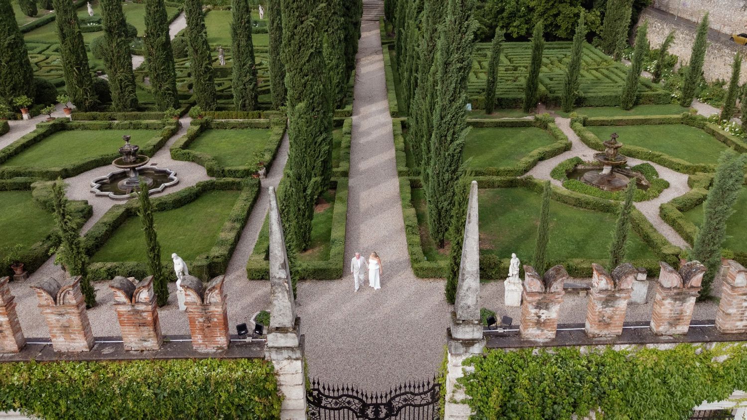 Aerial view of a formal Italian garden with cypress trees, fountains, and two people walking along a central stone pathway.