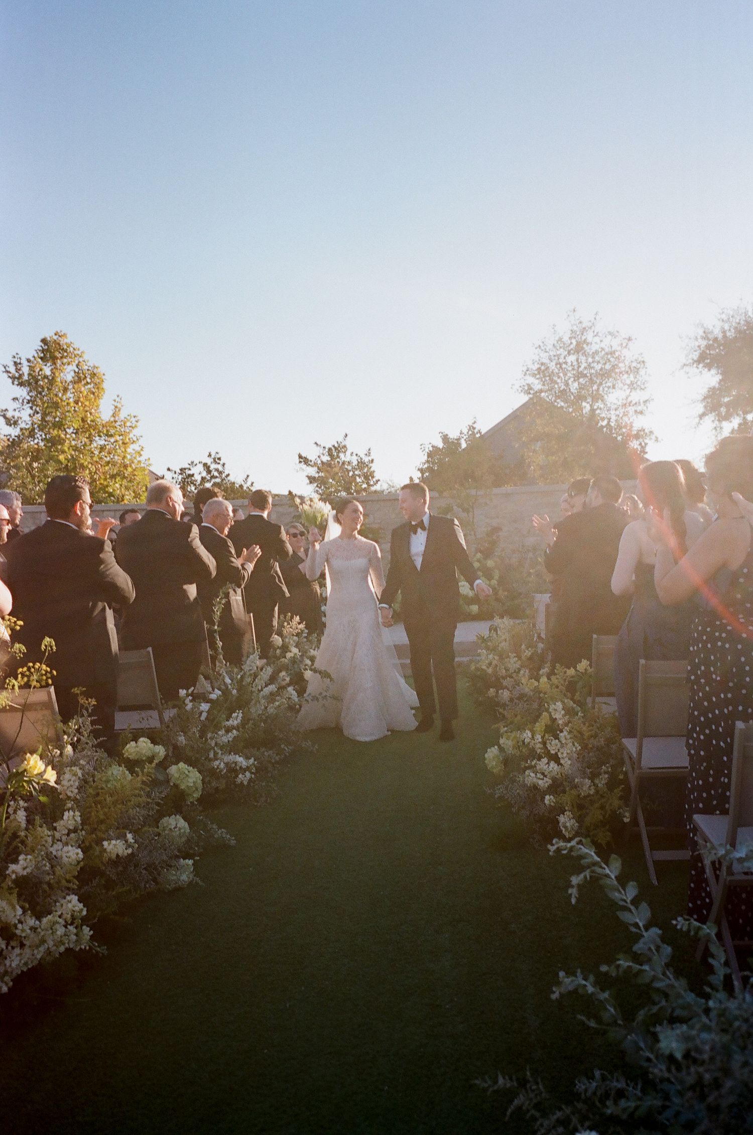 Wedding ceremony guests gathered outdoors with white floral decorations at sunset.