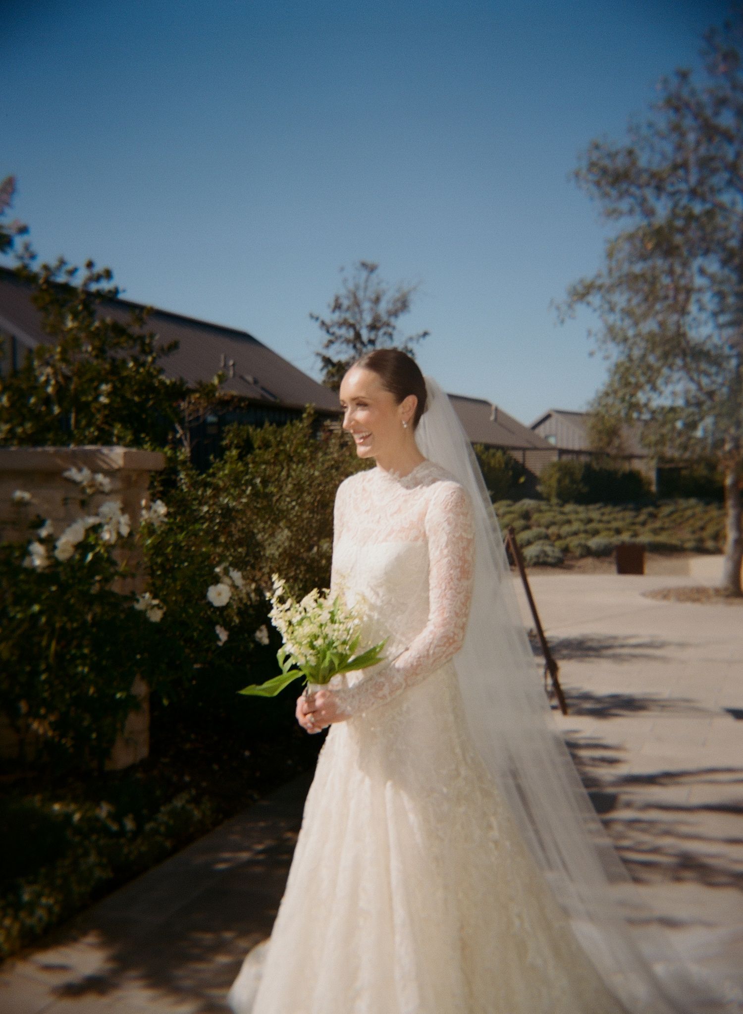 A bride in a long-sleeved lace gown poses outdoors holding a bouquet.