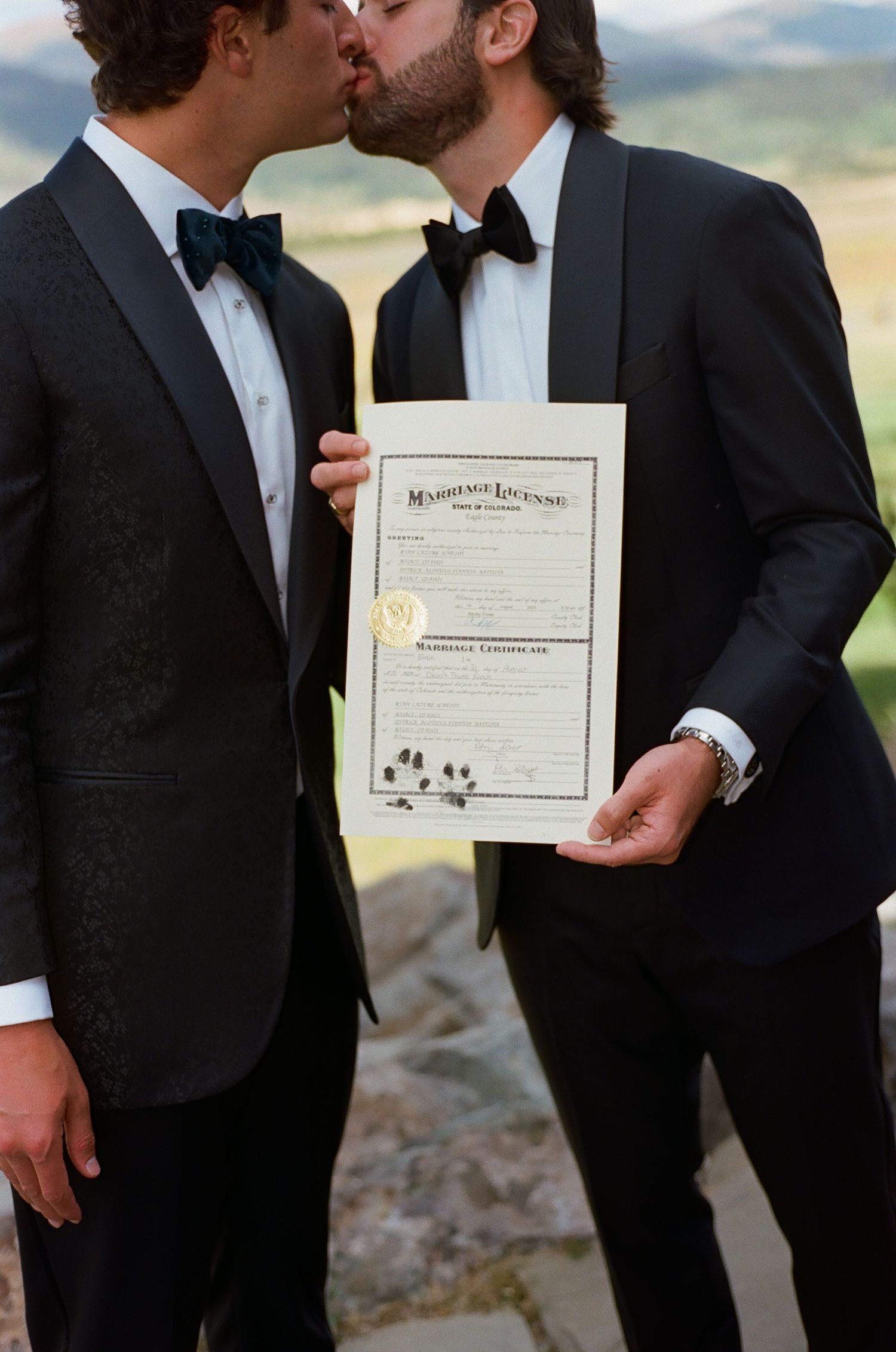 Groomsmen in black tuxedos holding a marriage certificate outdoors.