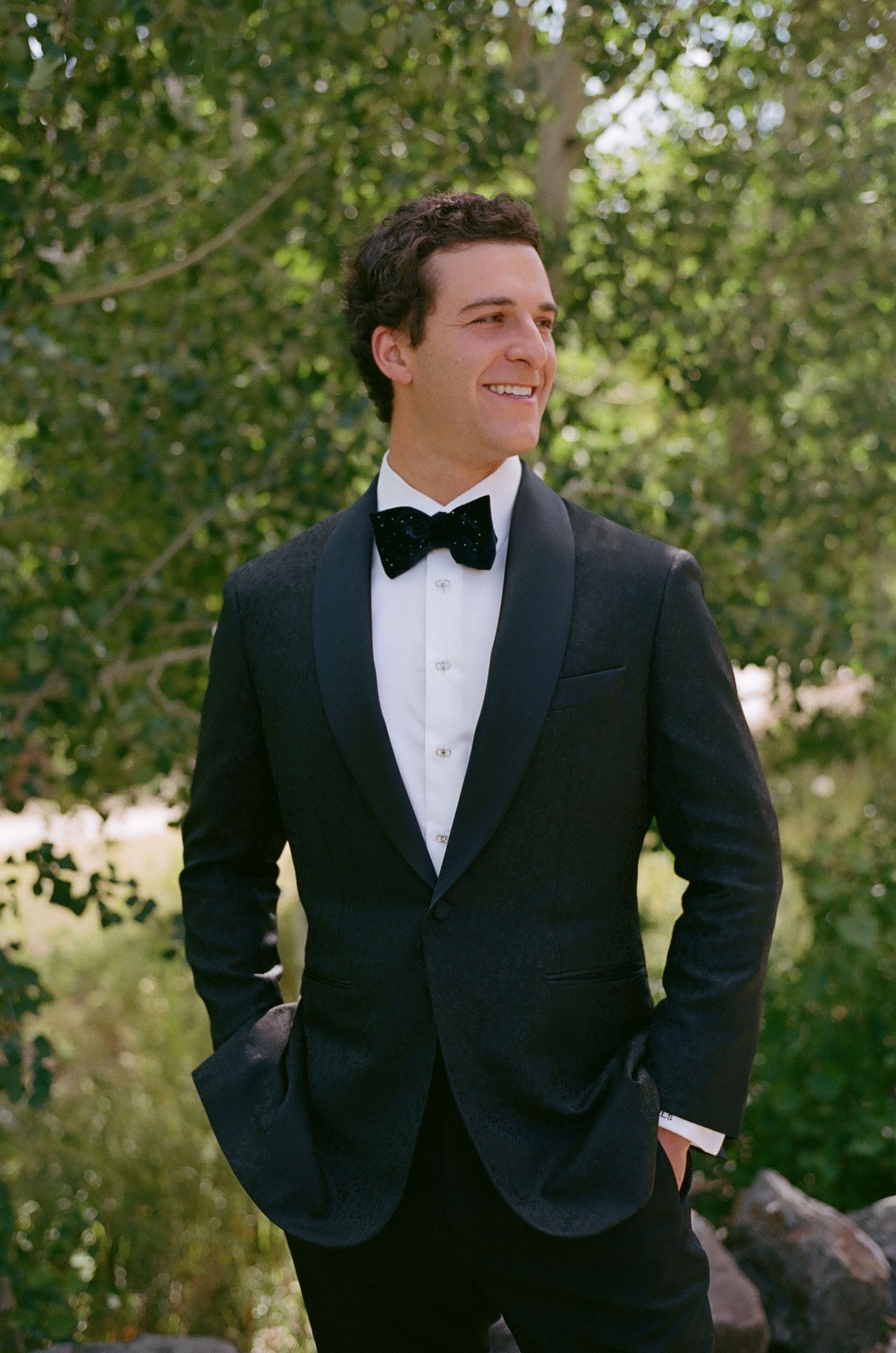 Person in formal black tuxedo and bowtie smiling outdoors with trees in background.