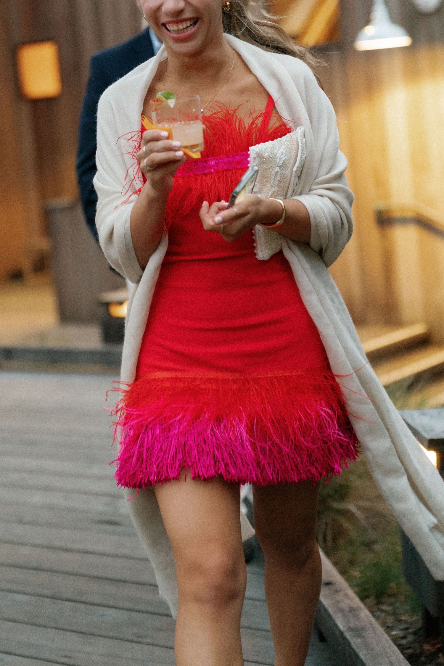 Wedding guest in red feathered dress and white cardigan holding drink on deck.