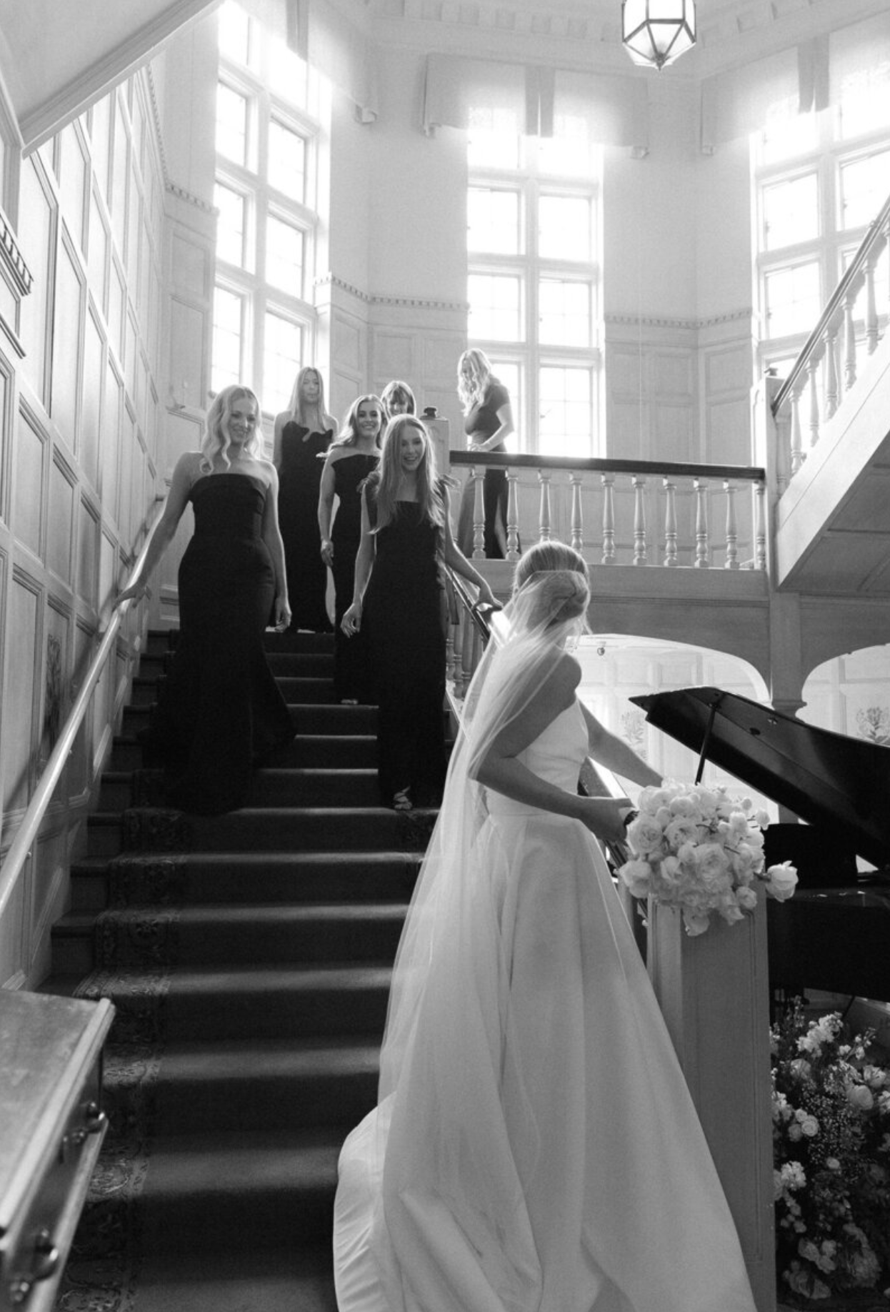 A bride in a white gown stands at the bottom of an elegant staircase while others in black dresses gather above.