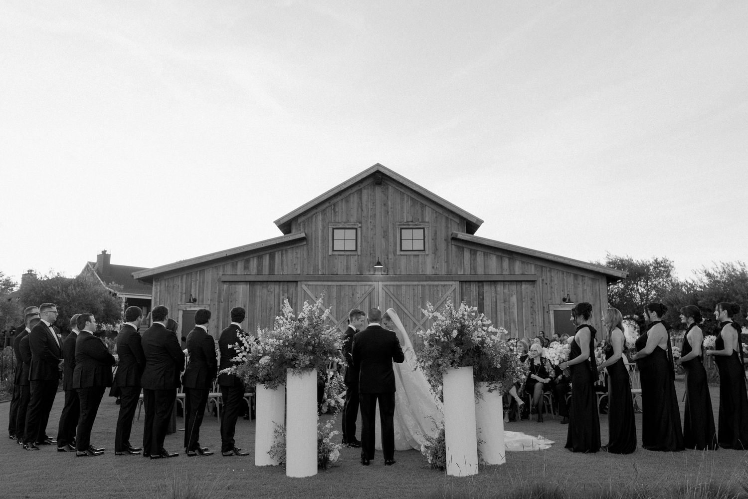 Wedding party lineup in front of a rustic barn with white floral pillars.
