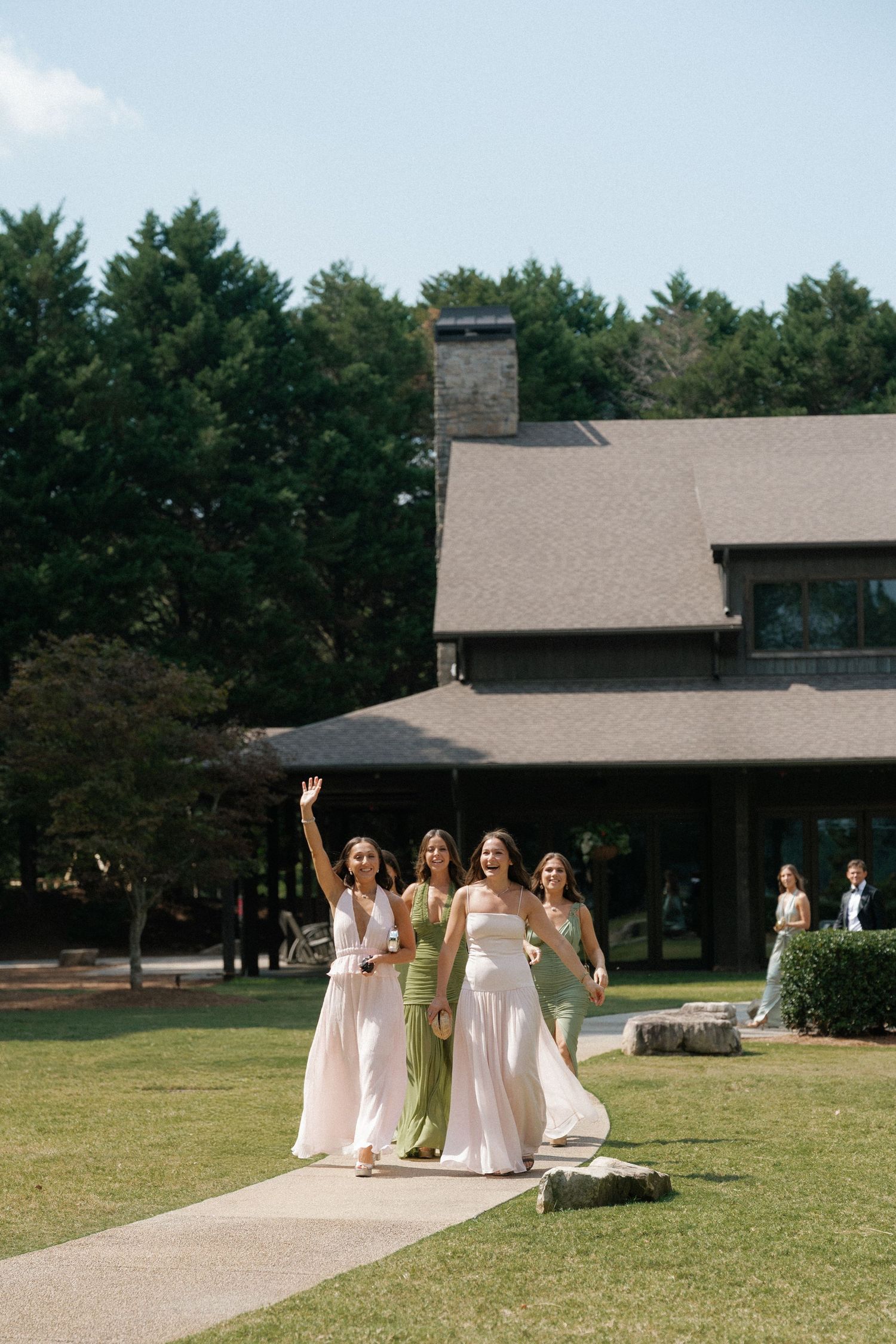 Bridesmaids in pink and green dresses walk together on a path leading to a rustic venue.