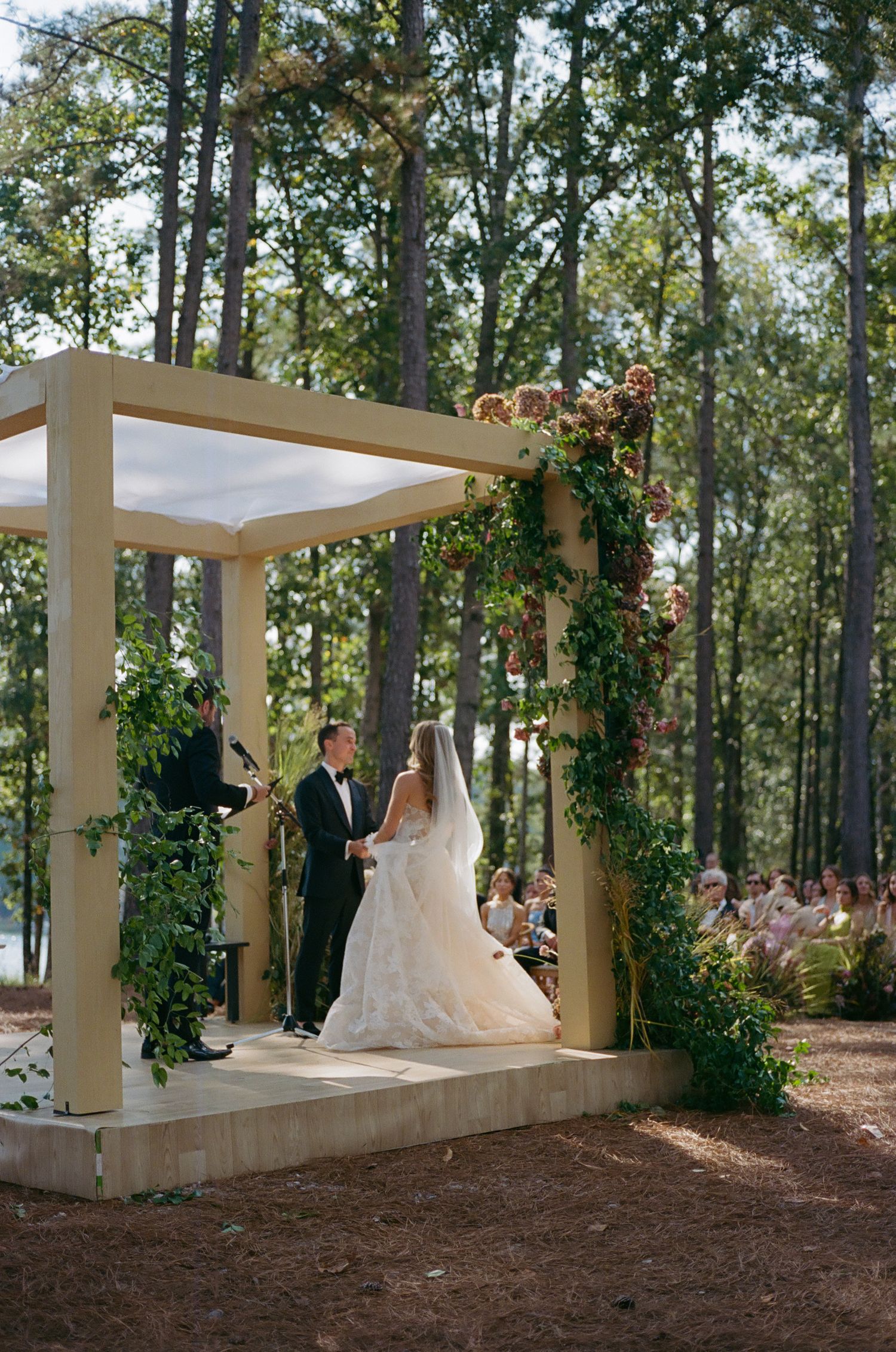 Elegant outdoor wedding ceremony setup with white pergola and green vine decorations by a lakeside forest.