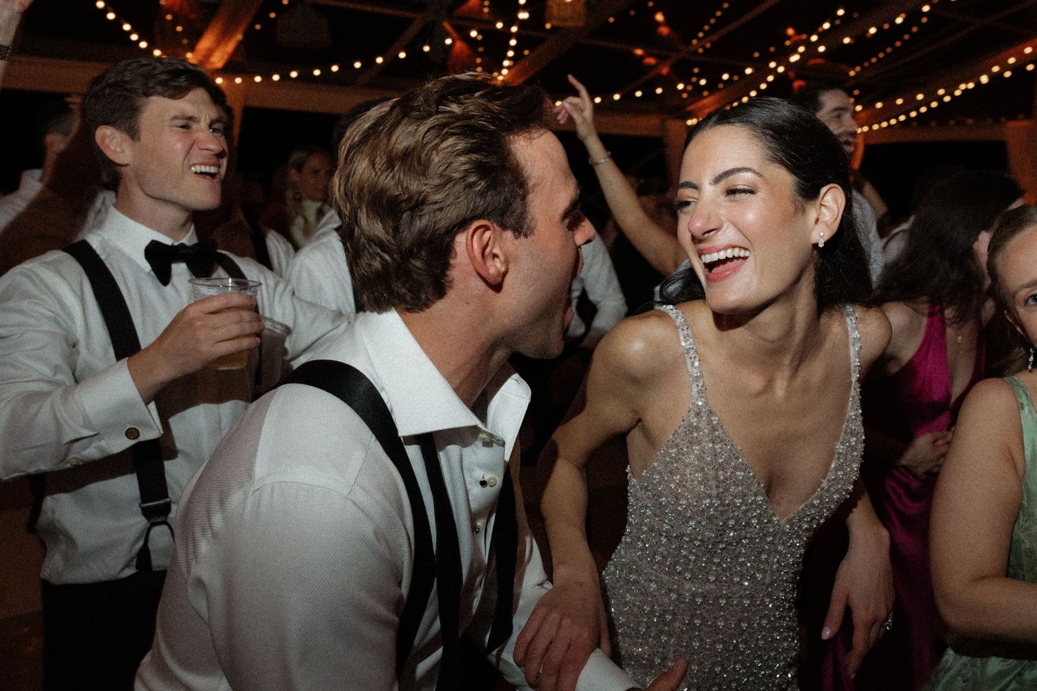 Joyful wedding guests dancing and laughing at lively wedding reception under warm lights.