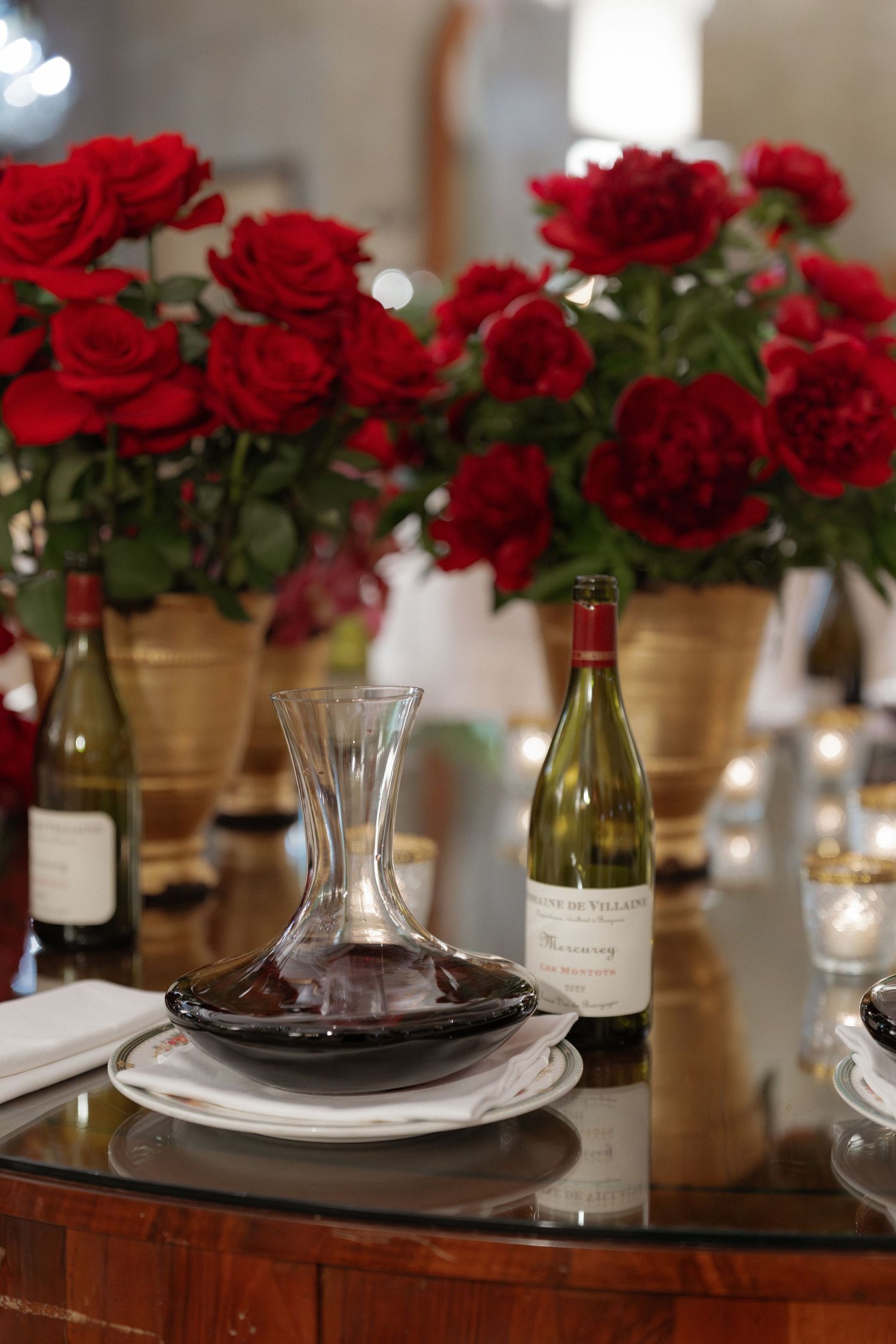 Wedding reception table with wine decanter, white wine bottle, and red roses in gold vases.