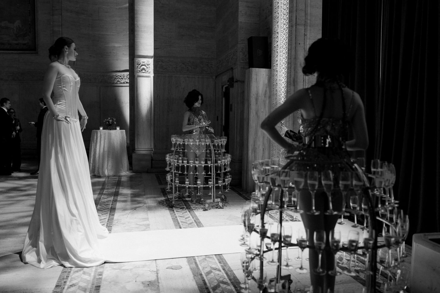 A black and white image of a woman in a long, flowing wedding dress standing in a grand, ornate room.