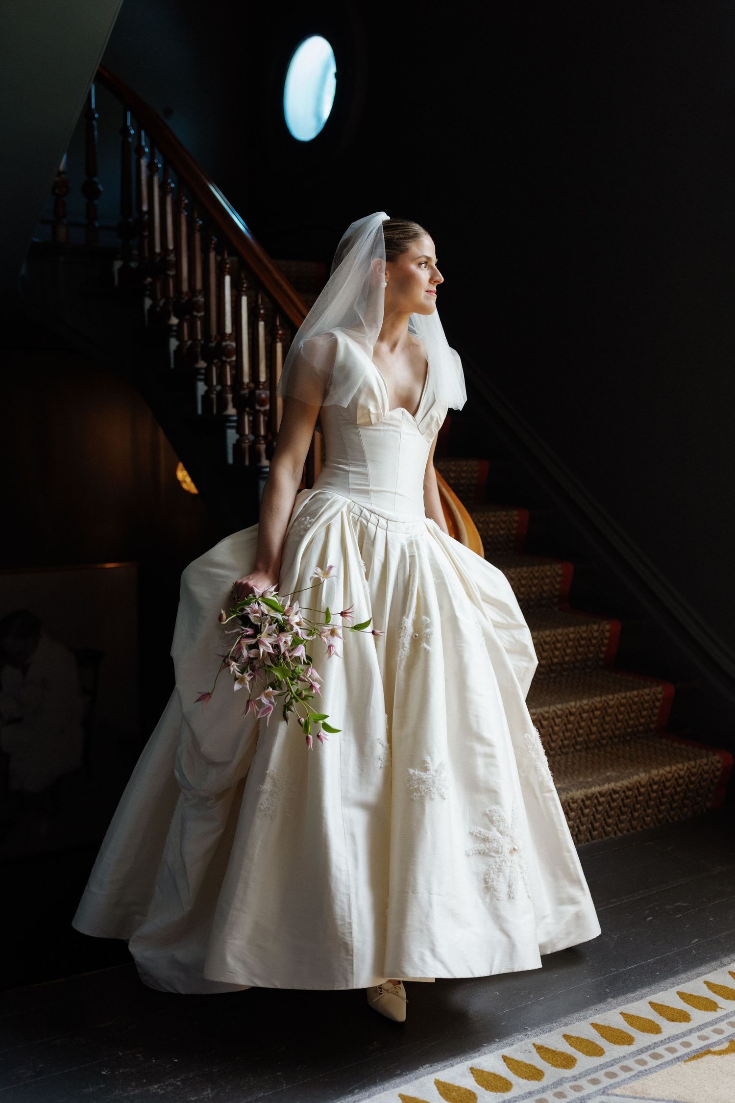 Elegant bride in a white ball gown with veil holds pink bouquet on staircase in dramatic lighting.