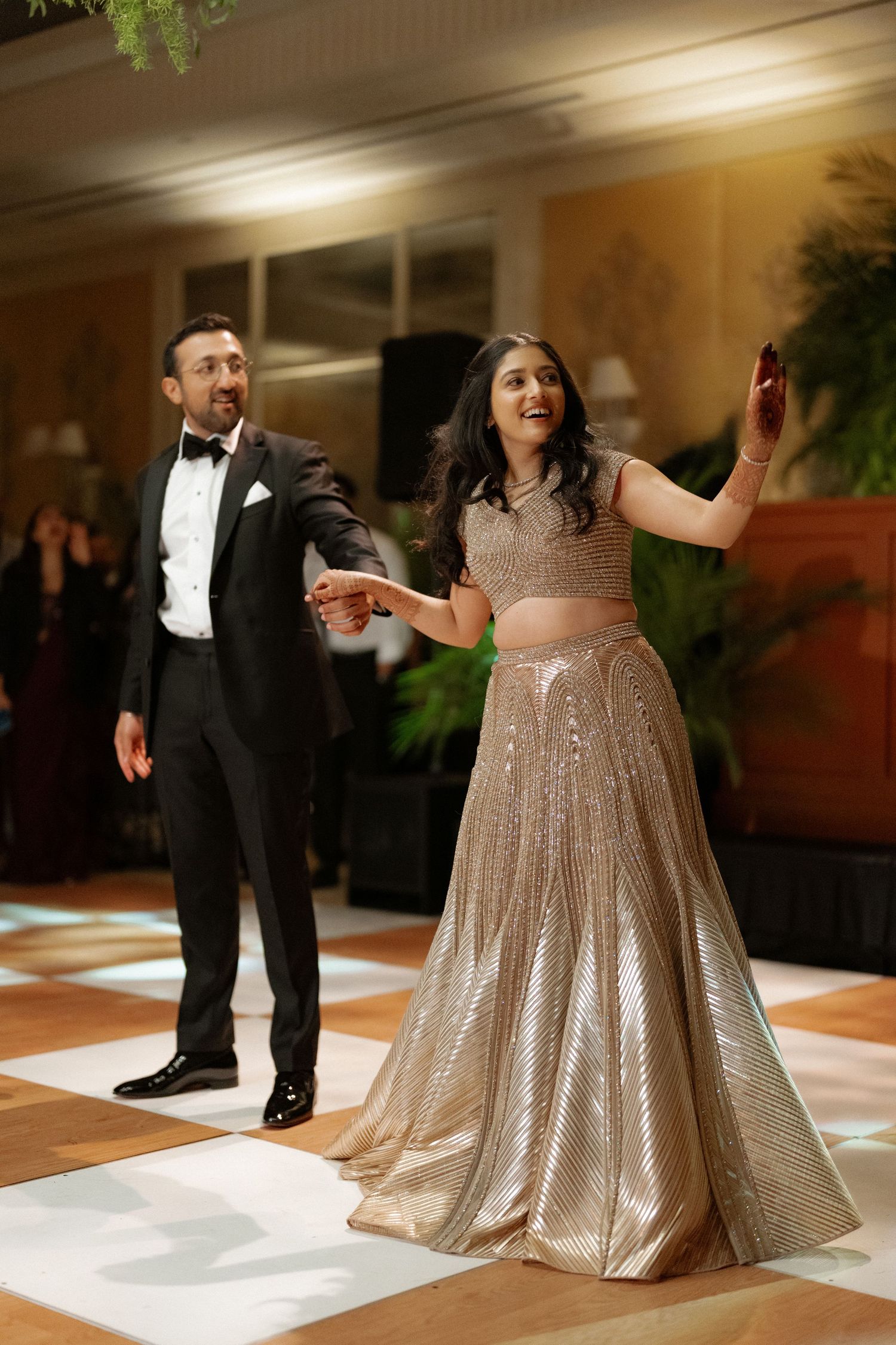 Couple dancing at wedding reception, man in tuxedo and woman in gold lehenga on dance floor.