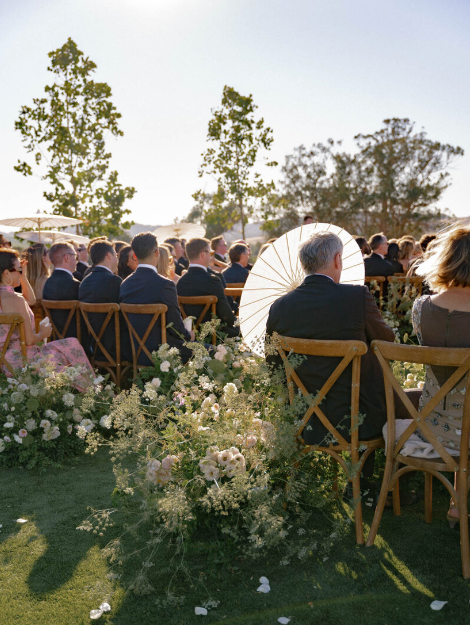 Wedding guests seated in wooden chairs watch an outdoor ceremony with white floral arrangements.