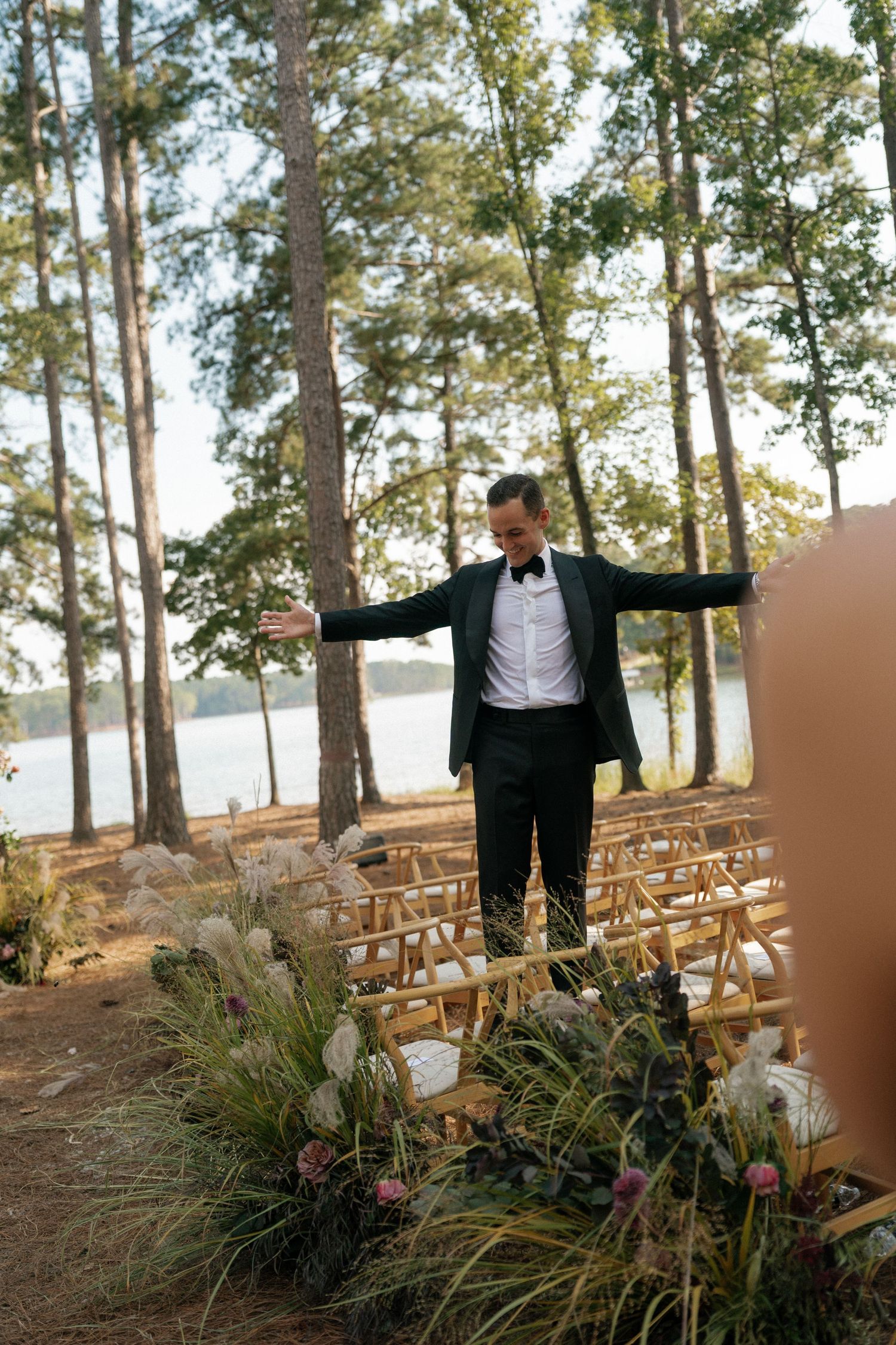 Wedding ceremony setup with wooden chairs and floral arrangements overlooking a lake among tall pine trees.