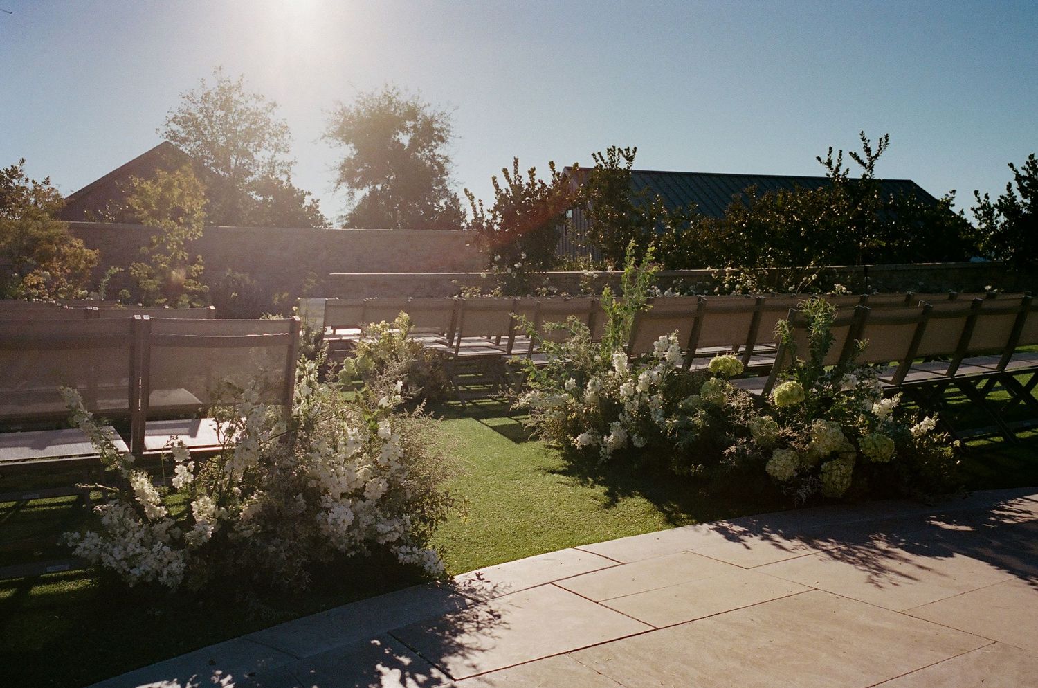 A sunny garden landscape with stone pathways and green lawns surrounded by buildings.