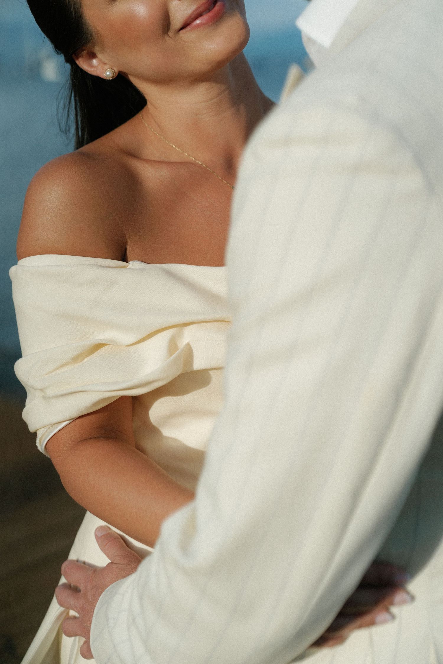 Intimate close-up of two people in cream-colored formal wear embracing by the water.