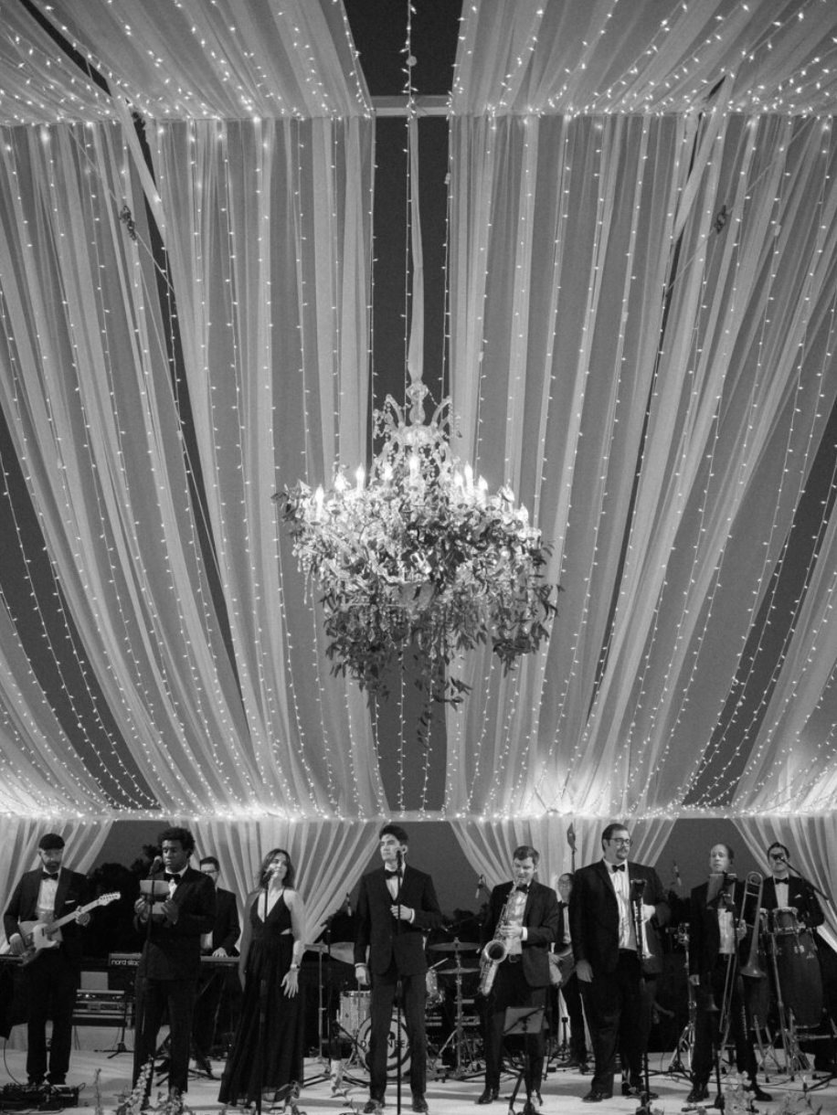 Black and white photo of a wedding band performing under draped fabric and crystal chandelier.