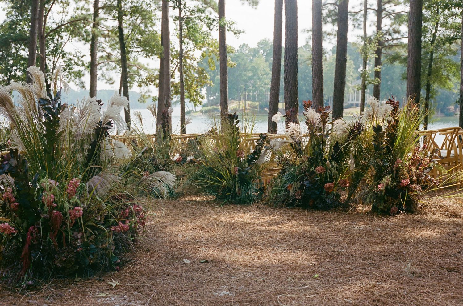 Natural outdoor wedding ceremony setup with rustic floral arrangements along a dirt path between pine trees.