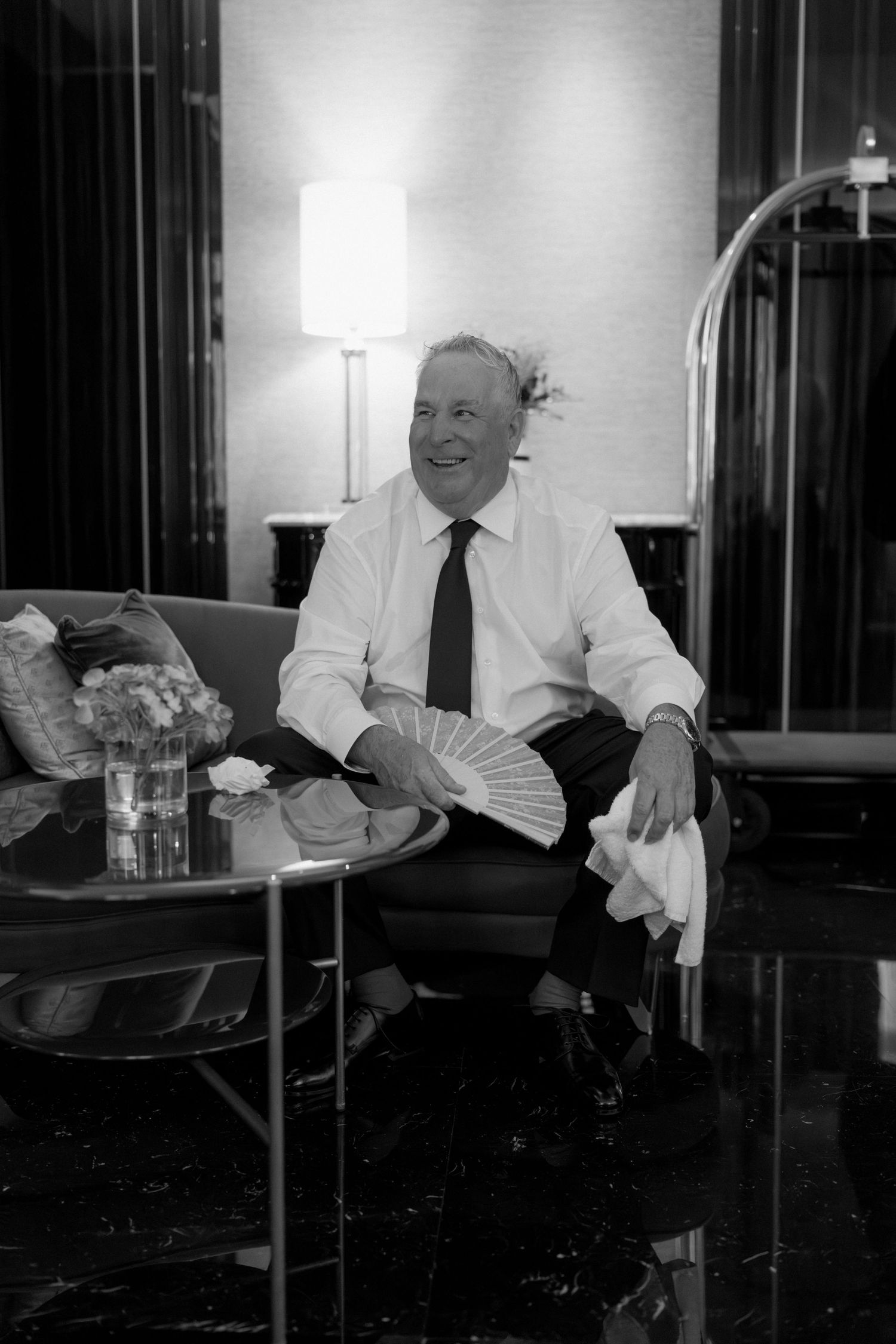 Smiling older man in white shirt and tie sits in luxury hotel lobby holding a hand fan in black and white.