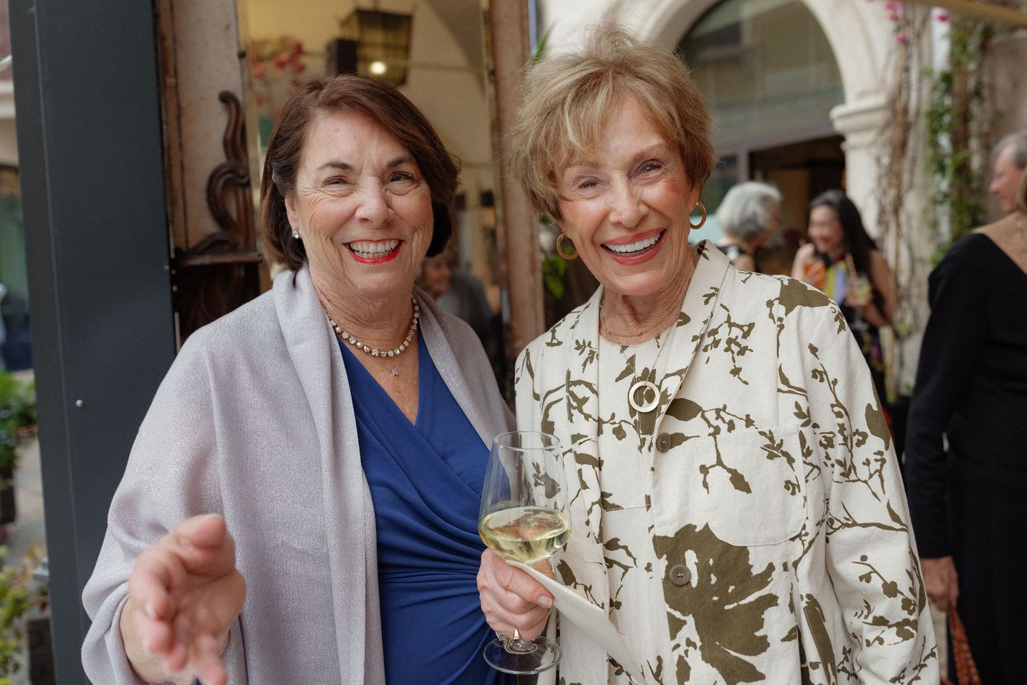 Two smiling women at a social event, one holding a wine glass, wearing elegant attire in a decorated venue.