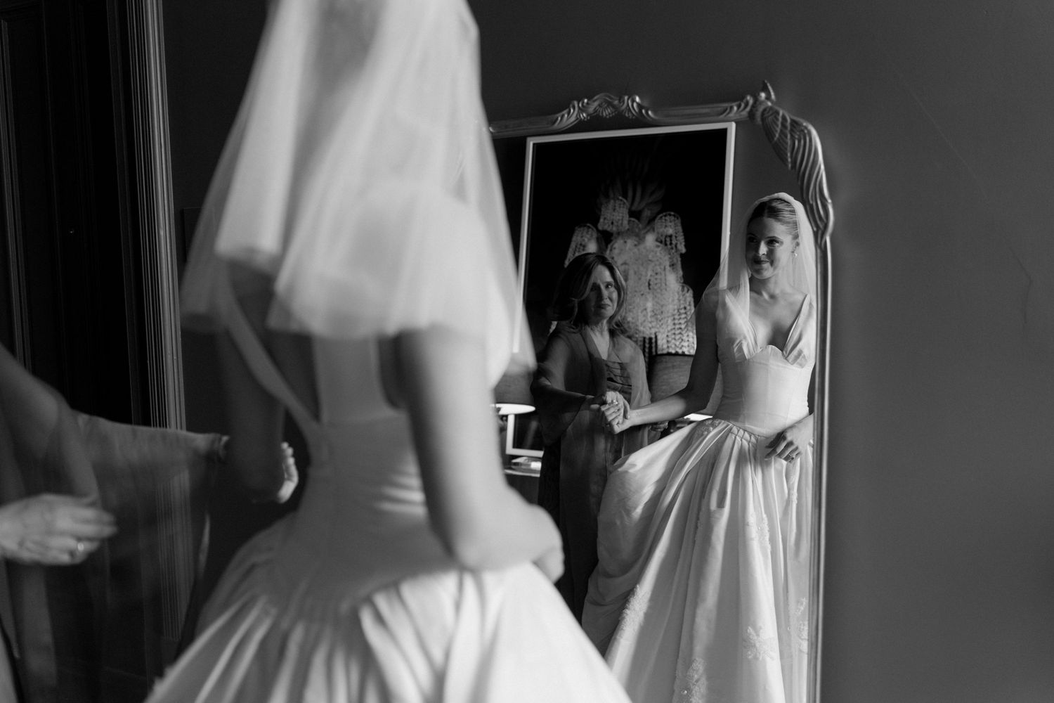 Black and white photo of a bride in a wedding gown preparing with bridesmaids, reflected in a vintage mirror.