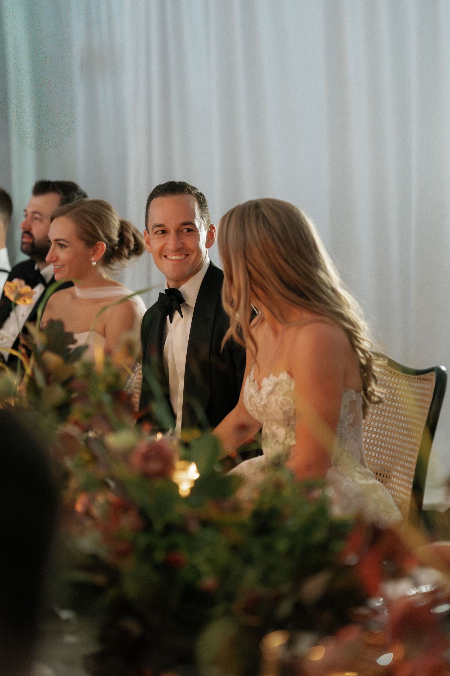 Wedding reception dinner table with elegant floral arrangements and white draping in the background.