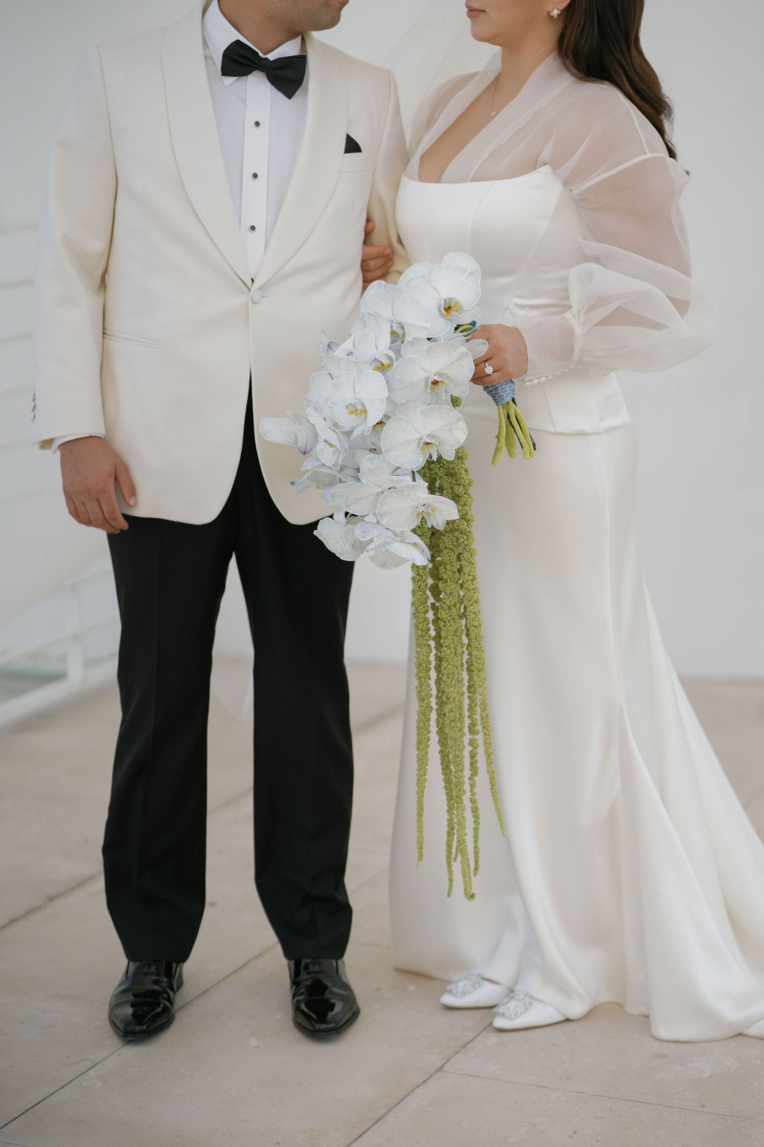 Wedding couple in white formal attire standing together with an orchid bouquet.