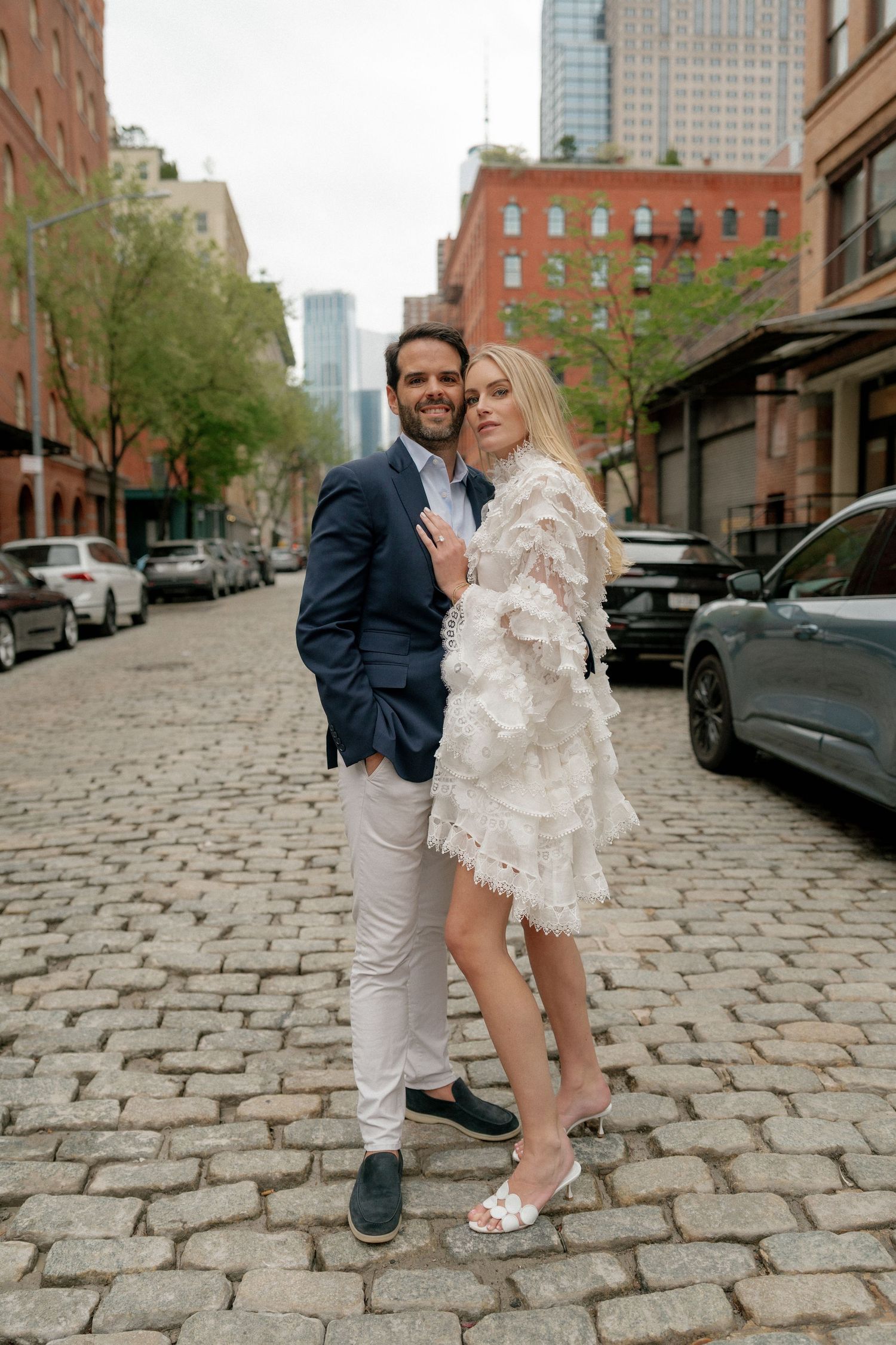 Couple poses playfully on a cobblestone street in an urban setting with brick buildings and city skyline in background.