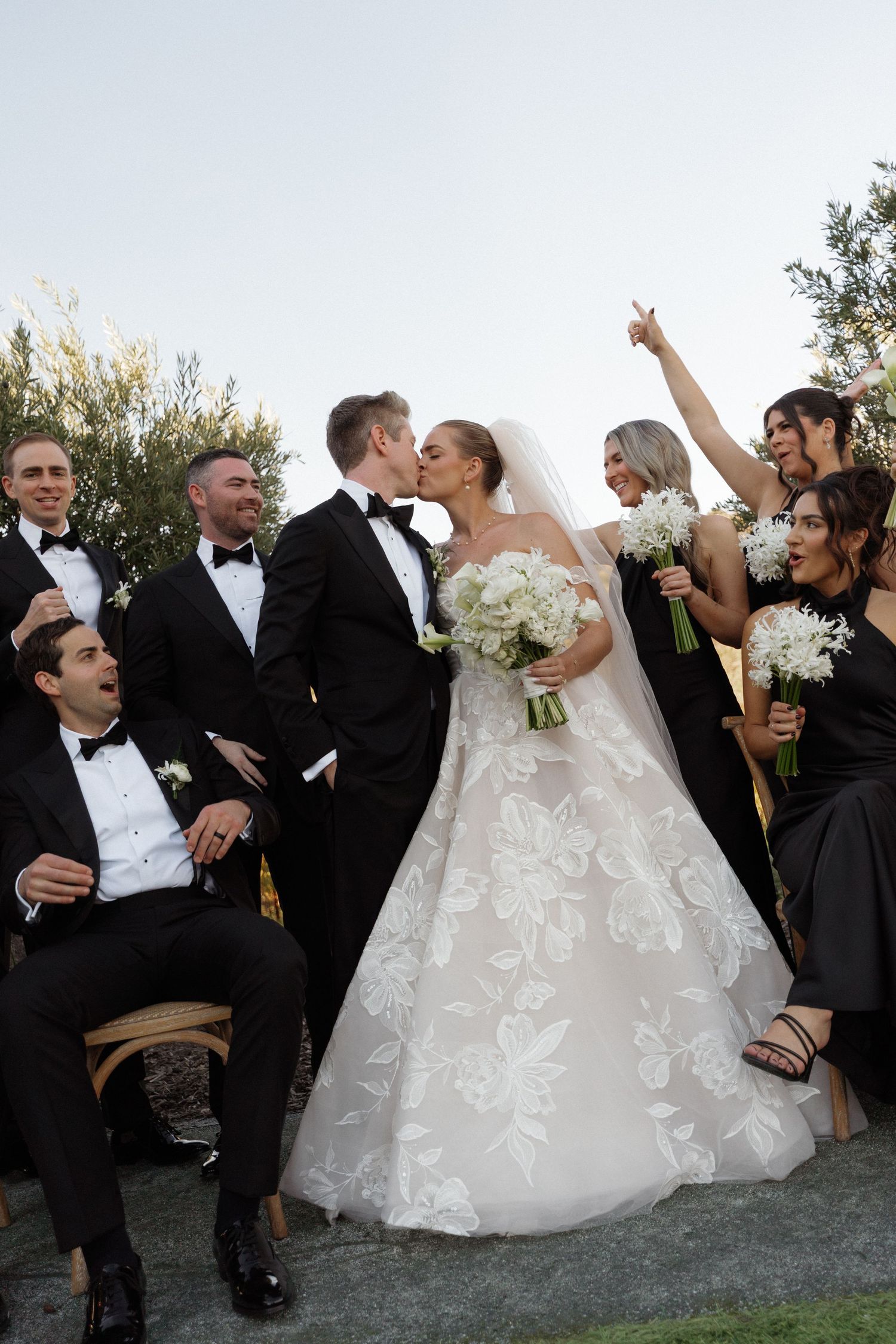 Wedding party in formal attire celebrates with newlyweds outdoors.