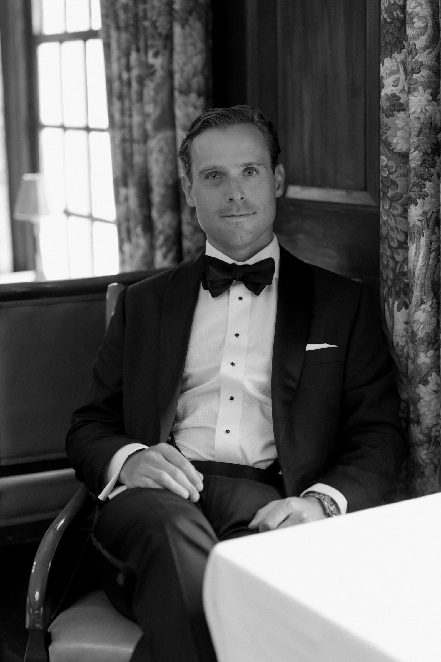 Elegant man in black tuxedo with bow tie seated at a table in a formal dining room, black and white photo.