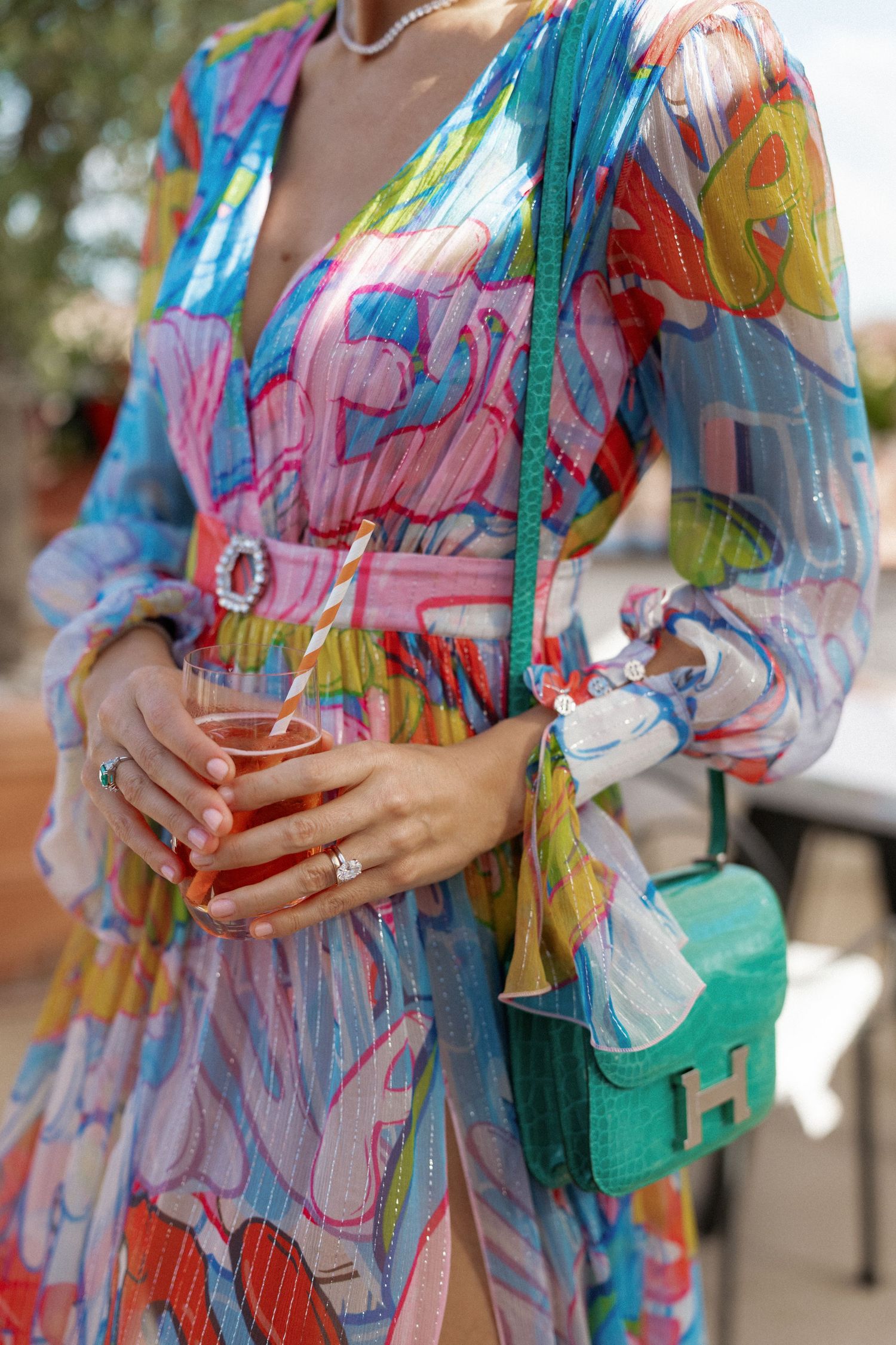 Woman in colorful printed maxi dress with green Hermes crossbody bag and diamond ring holding a drink.