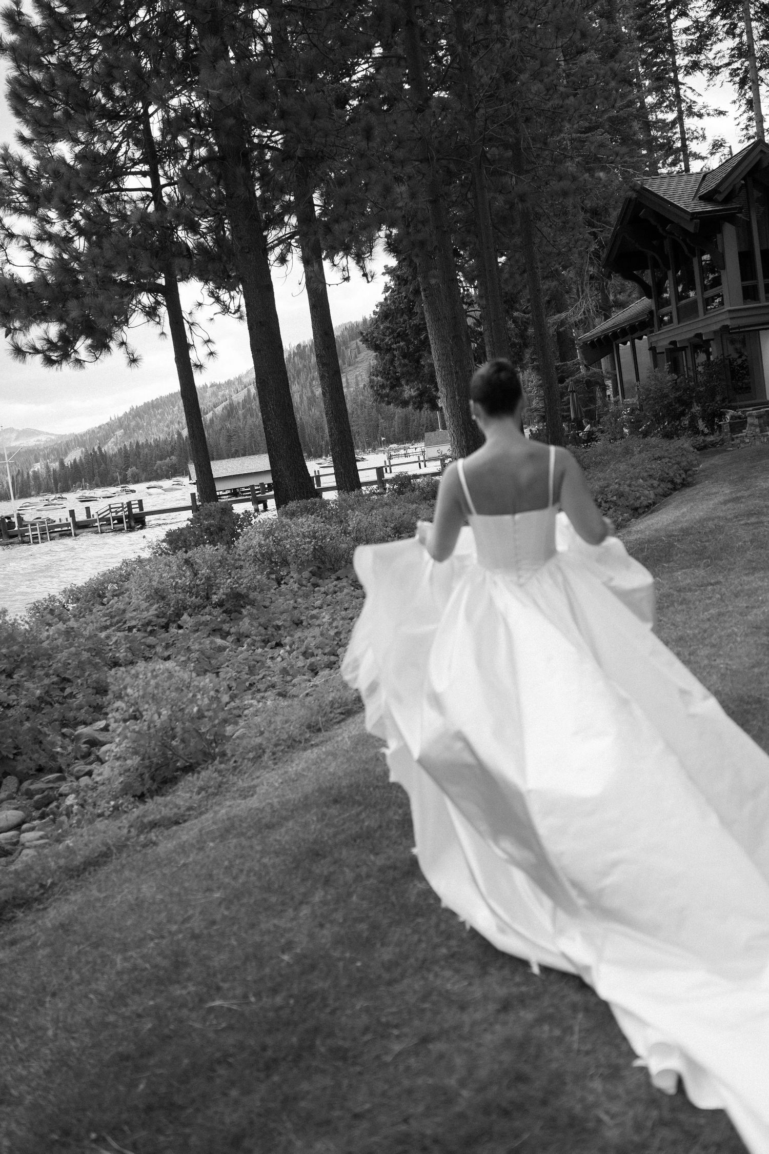 White wedding dress train flows across grassy hillside with pine trees in background.