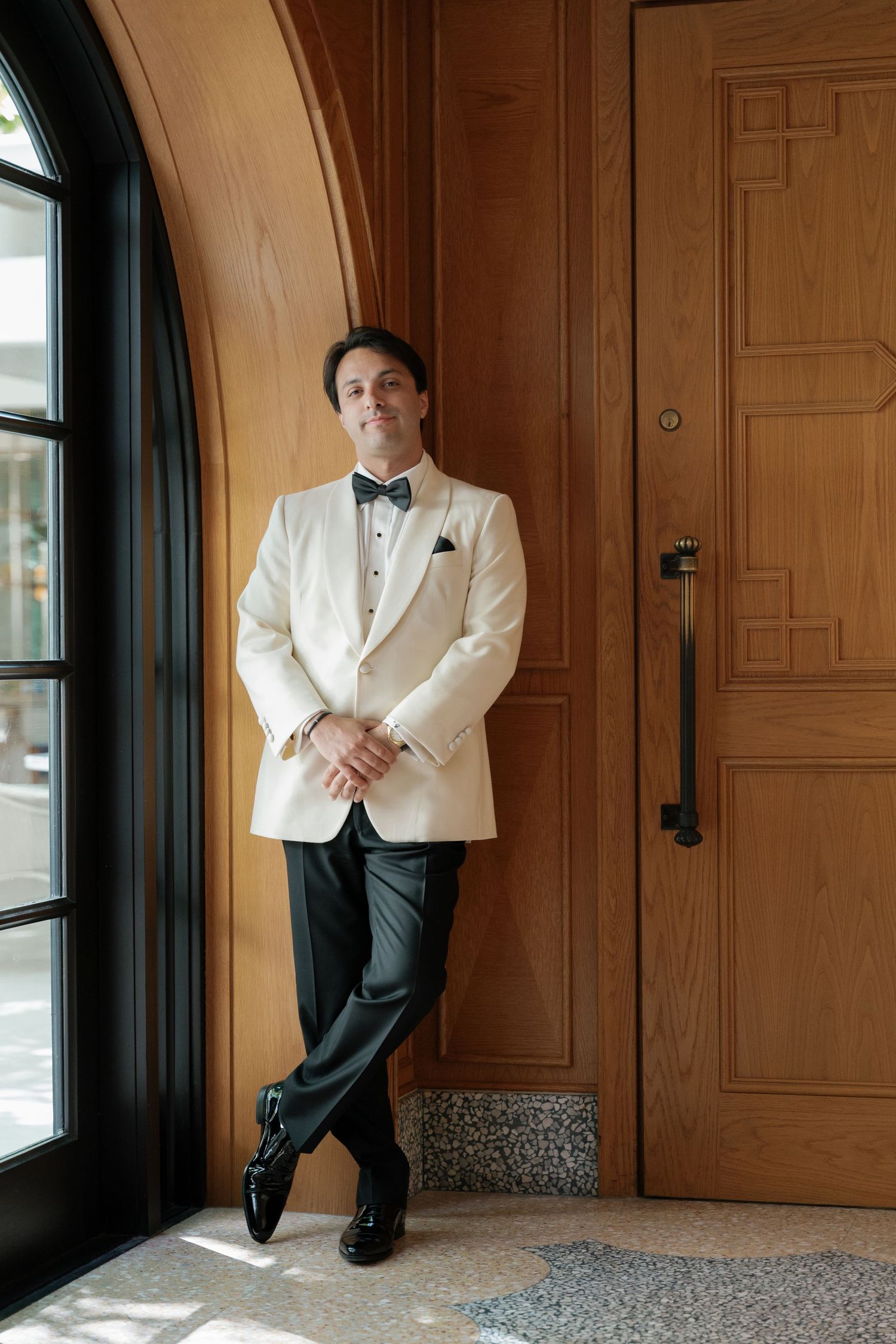 A groom in a white dinner jacket and bow tie poses casually by a window in an elegant venue.