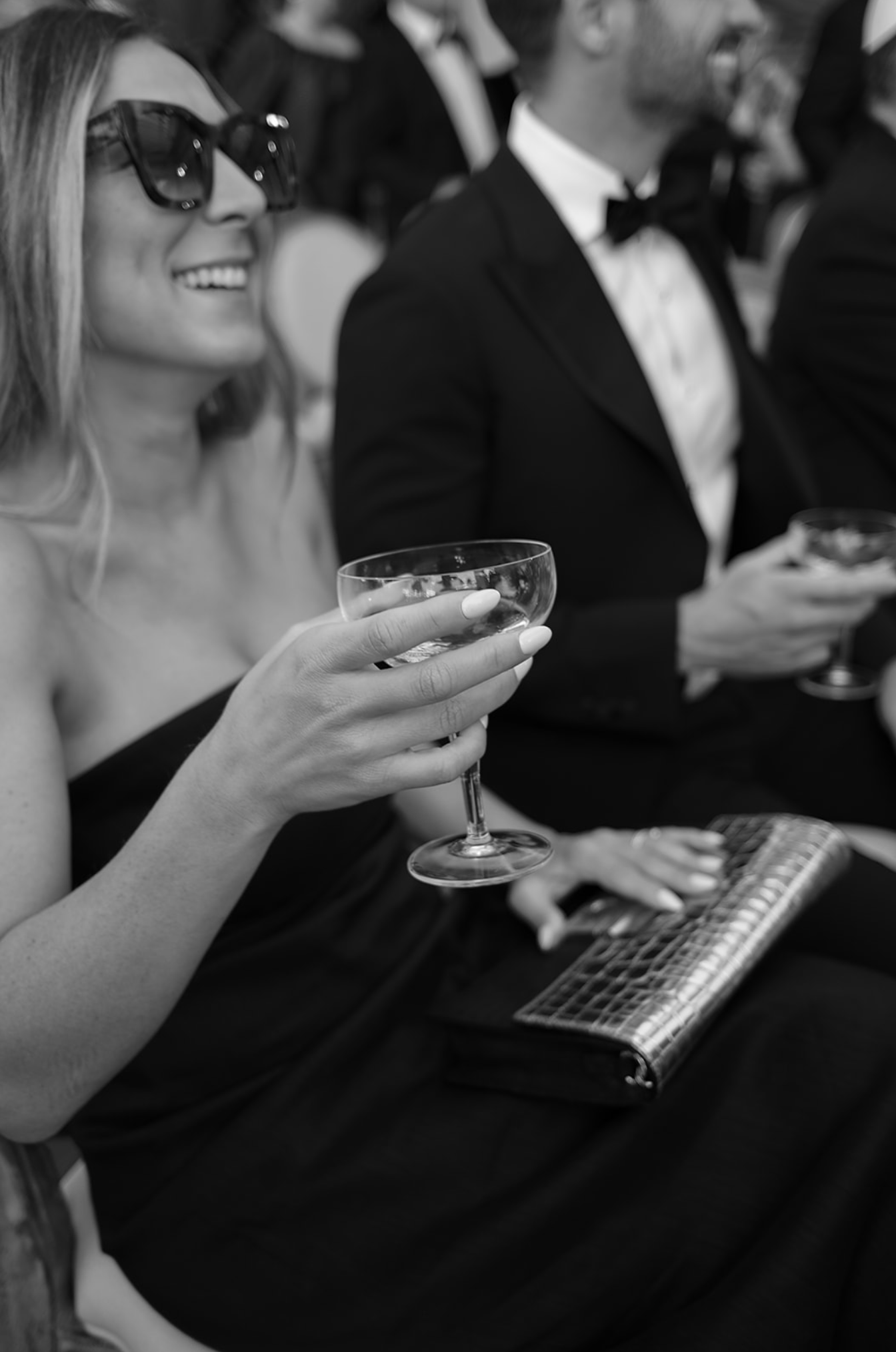 A guest in sunglasses holds a cocktail glass at an elegant event with a formal dress code.