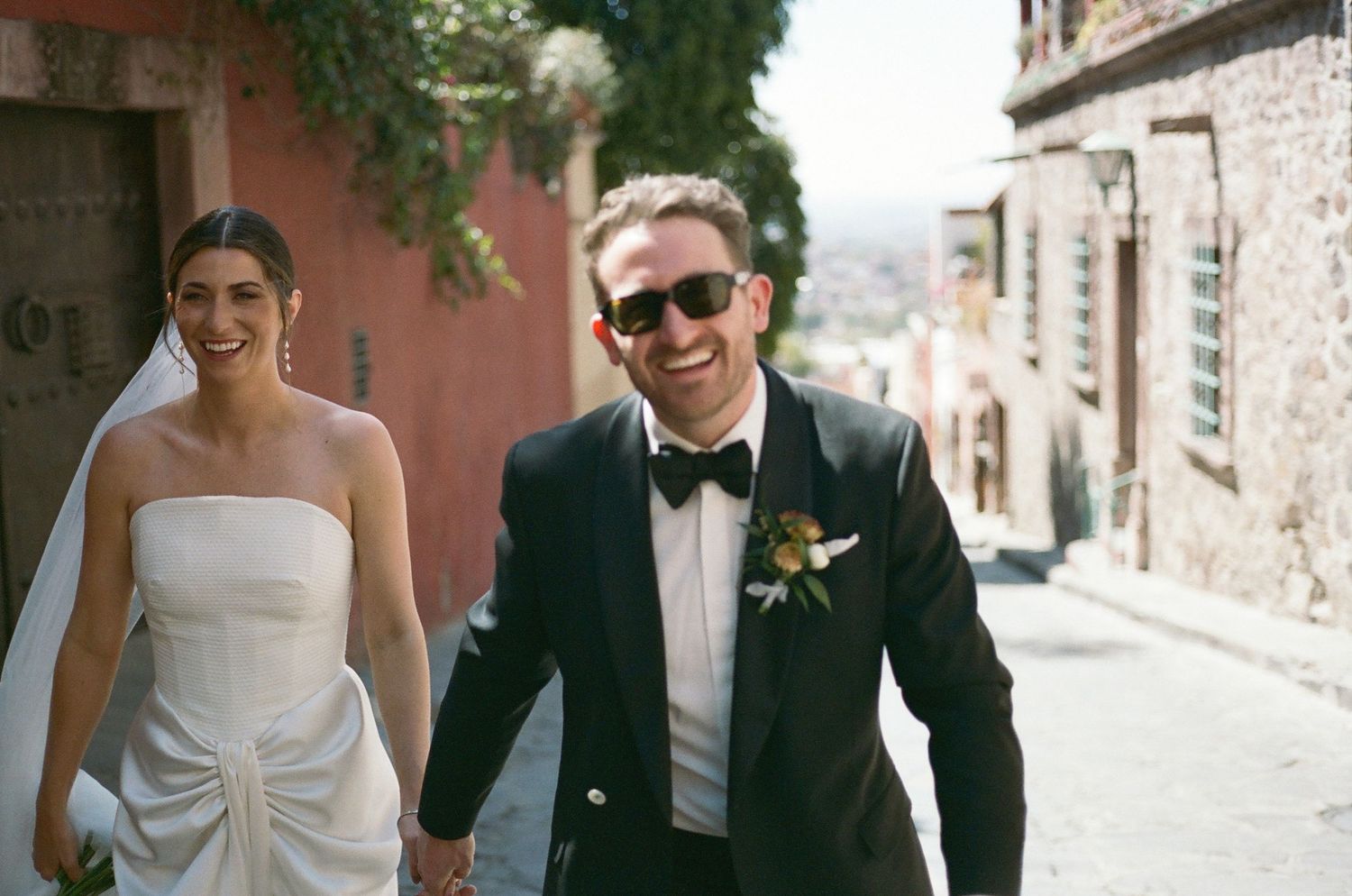 Wedding couple walking down sunny cobblestone street with pink colonial walls.