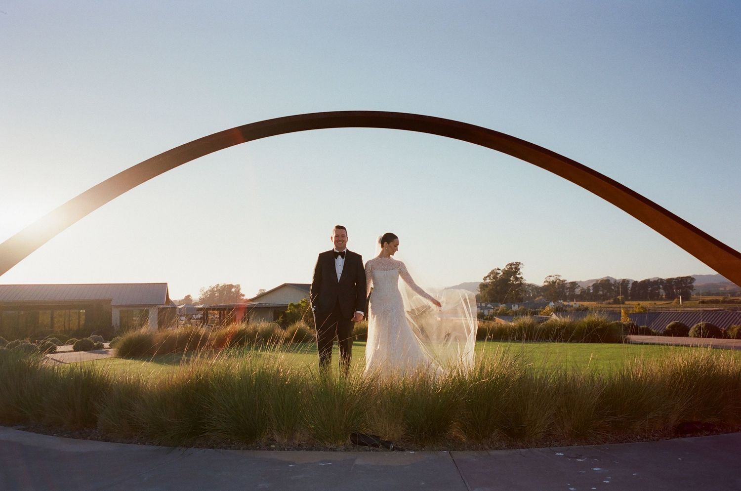 A couple in wedding attire stands beneath a curved arch at sunset on a grassy lawn.