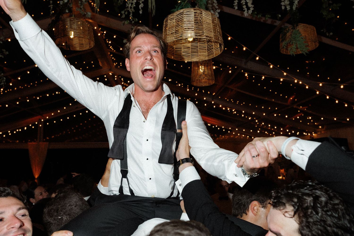 Excited groom in white shirt and suspenders crowd surfing at a lively wedding reception celebration.