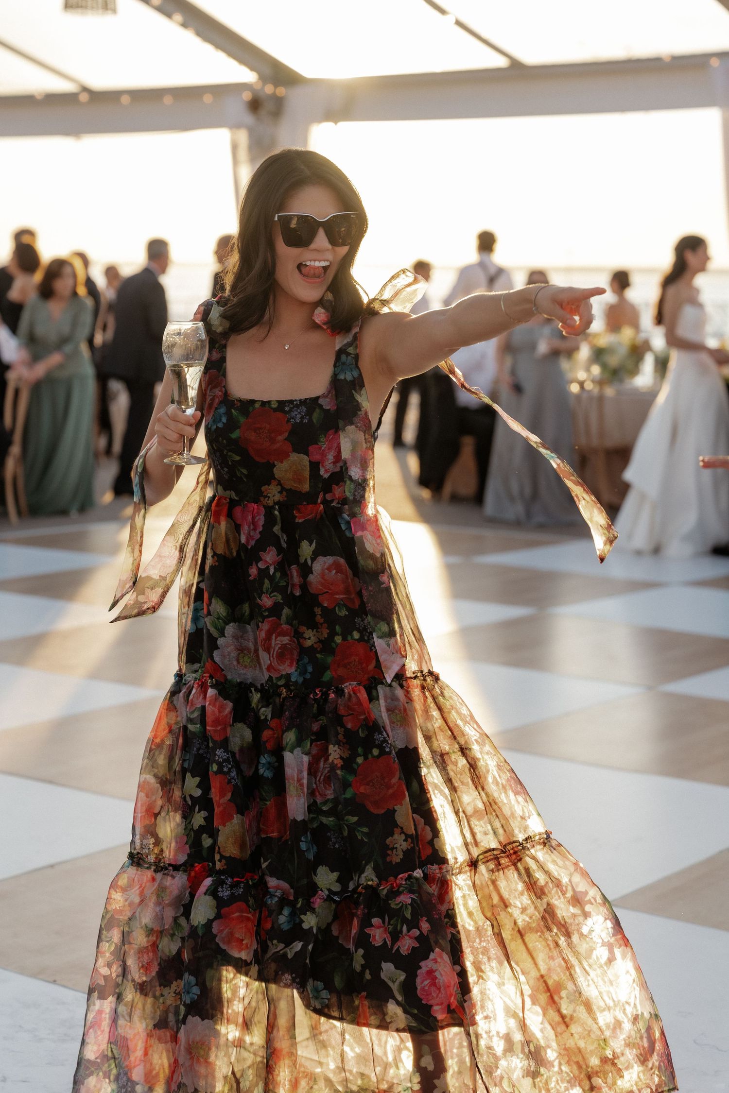Woman in floral dress dancing and pointing at a beachside wedding reception at sunset.