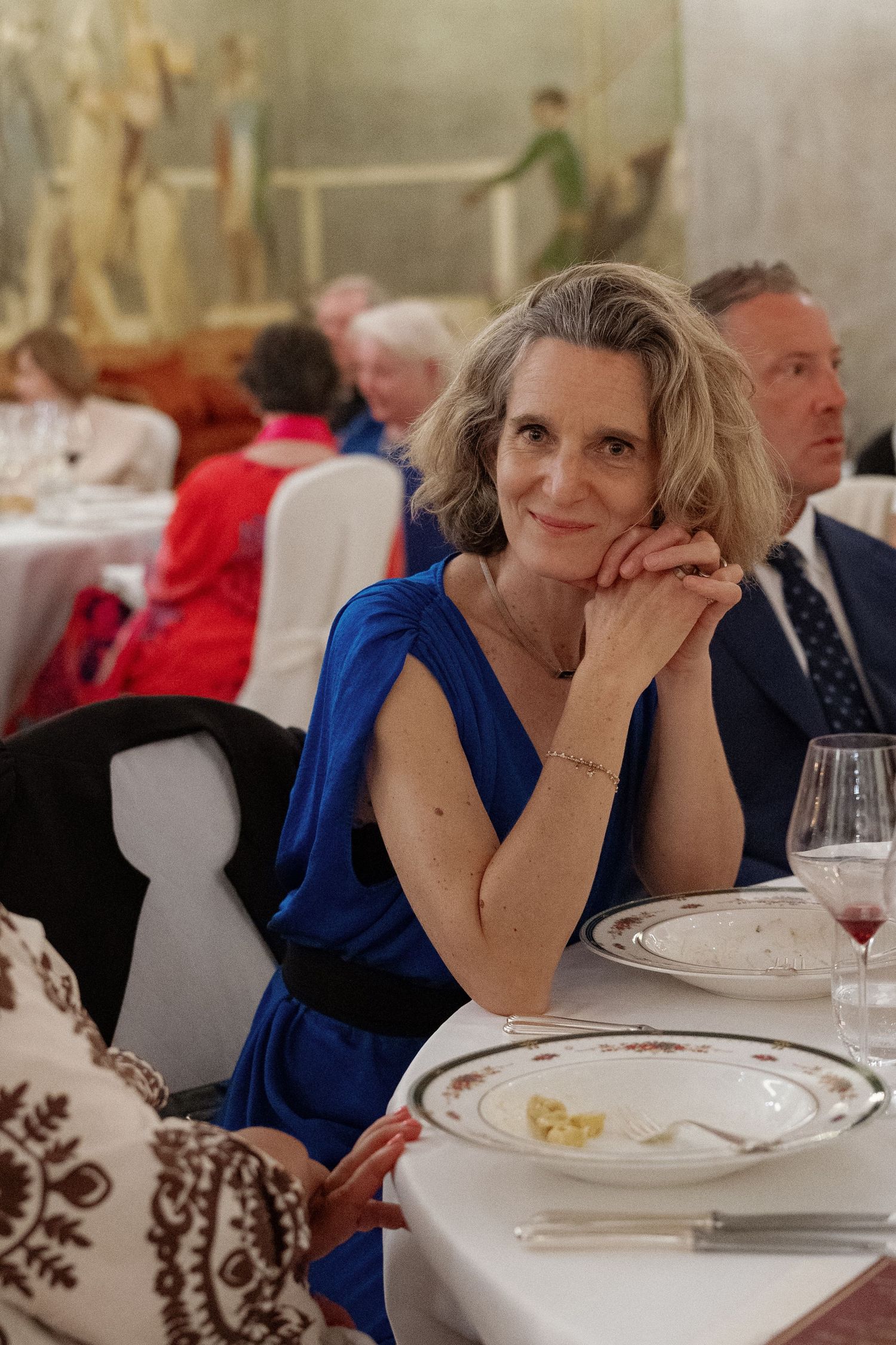 Elegant woman in blue dress seated at formal dinner table, smiling gracefully with hands clasped.
