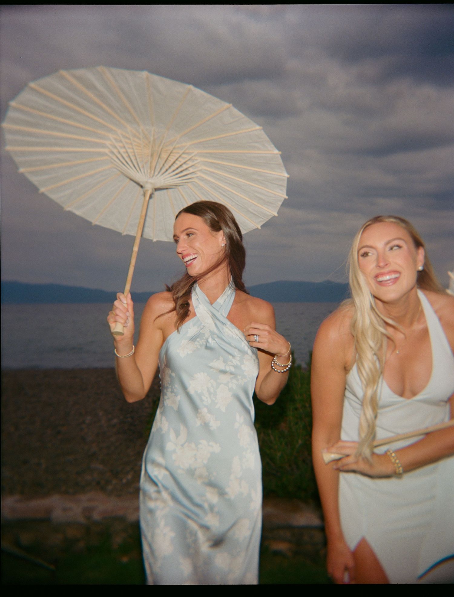 Two women in light dresses, one holding a decorative parasol outdoors.