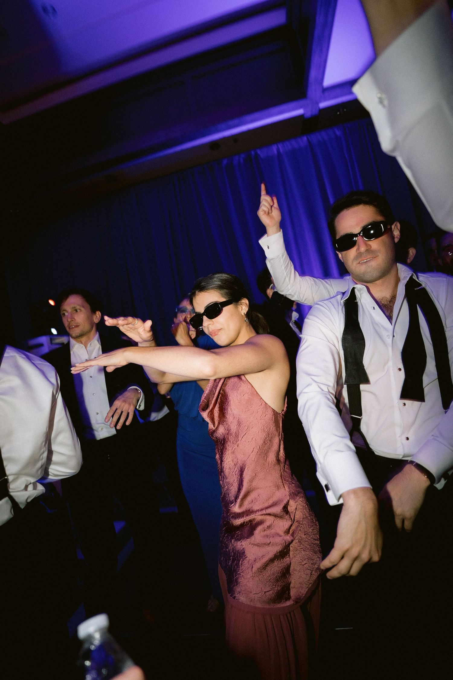Wedding reception dance floor with guests wearing sunglasses and dancing under purple lighting.